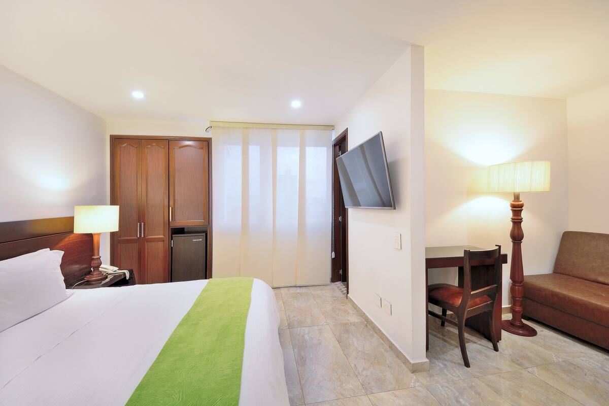 A hotel room with a bed, a desk, a chair, a sofa, a TV, and a lamp.