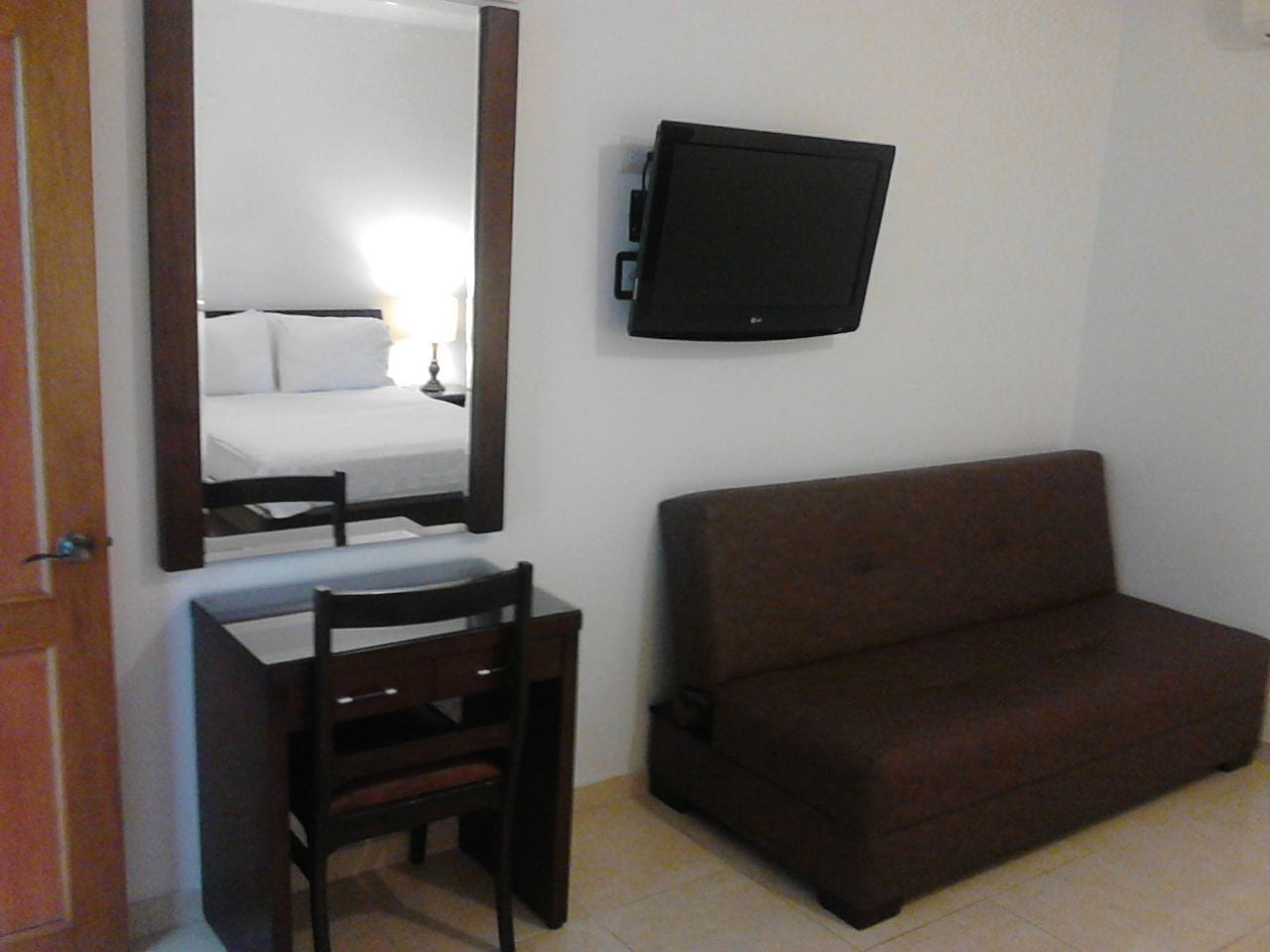 A room with a bed, a desk with a chair, a brown sofa, and a mounted television.
