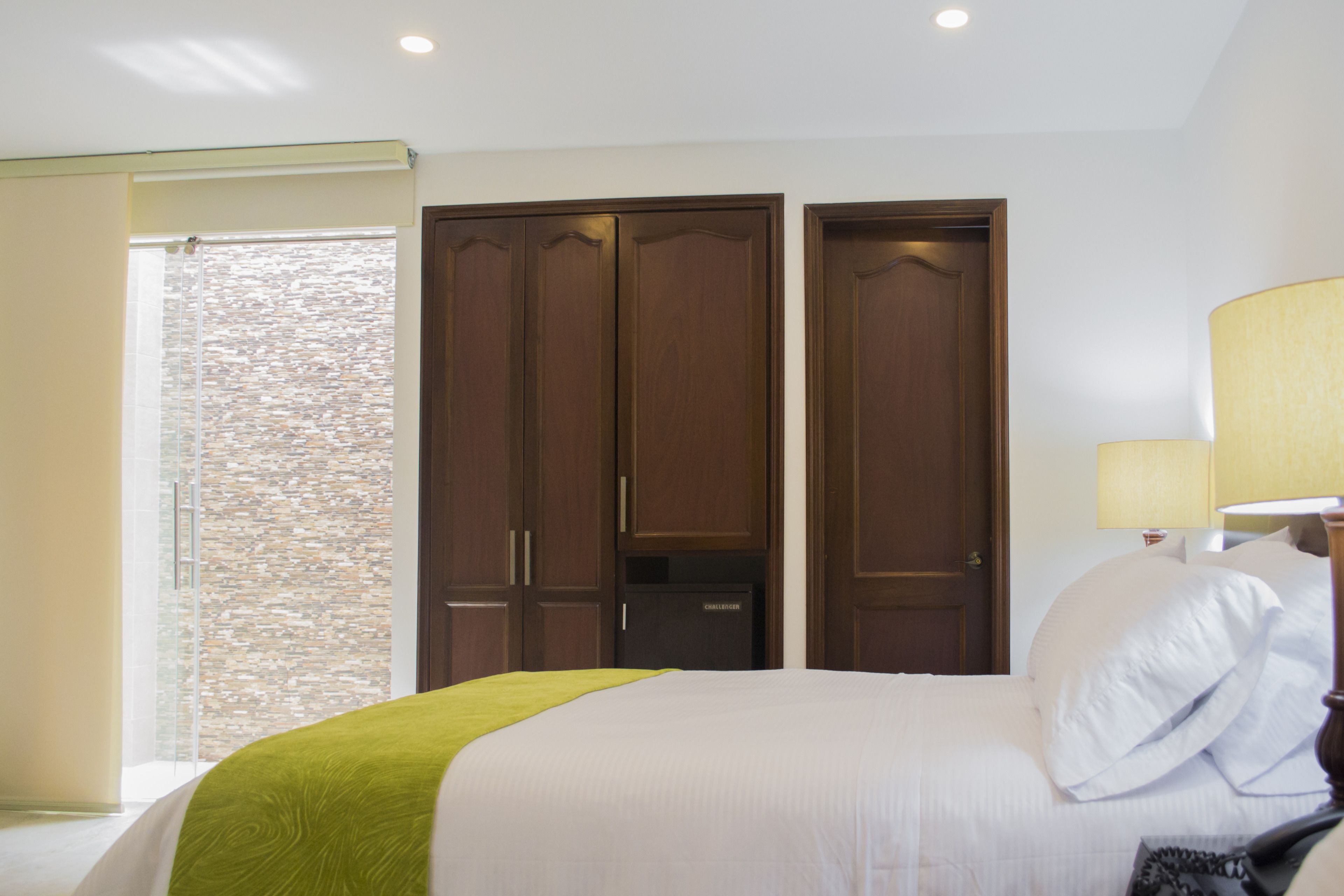 A hotel room with a bed, a wardrobe, a lamp, and a window with a view of a brick wall.