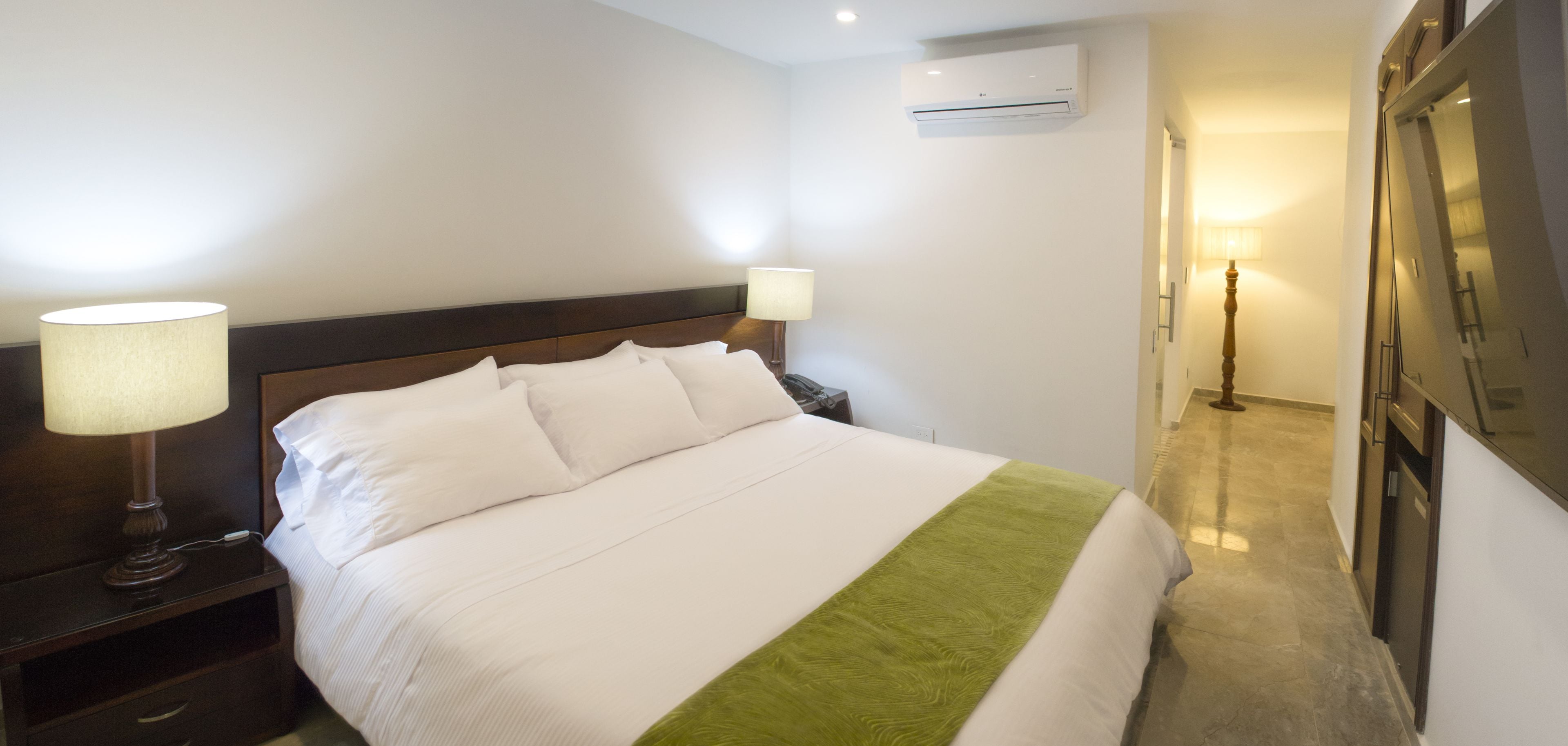 Ein Hotelzimmer mit einem ordentlich bezogenen Bett, Nachttischlampen, einem Nachttisch, einer Klimaanlage und einem Spiegel an der Wand.