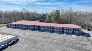 A large, single-story building with a red roof and multiple red doors, surrounded by a parking lot and a forested area. - Americas Best Value Inn Stearns (Stearns)