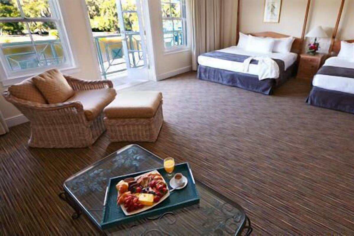 Camera d'albergo con due letti, una sedia in vimini, un tavolo da pranzo con un vassoio di frutta e vista sull'esterno.