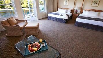 A hotel room with two beds, a wicker chair, a dining table with a fruit platter, and a view of the outdoors.