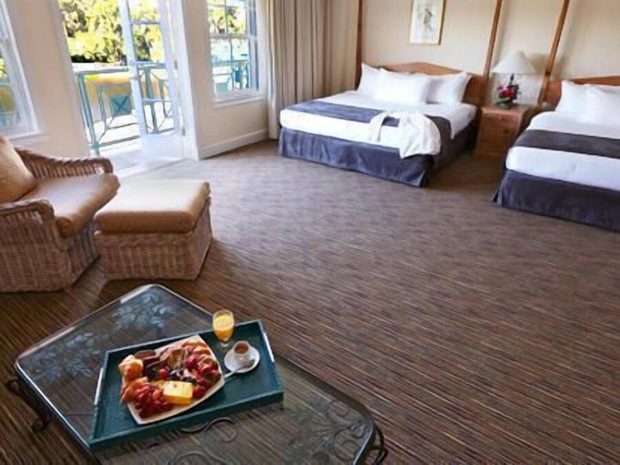 A hotel room with two beds, a wicker chair, a dining table with a fruit platter, and a view of the outdoors.