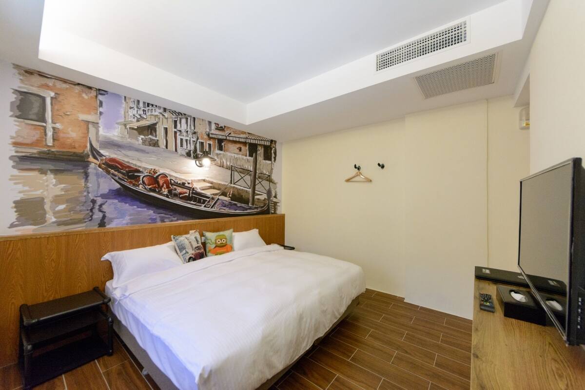 Une chambre d’hôtel avec un lit, une télévision, une petite table et une fresque murale représentant un paysage de canal.