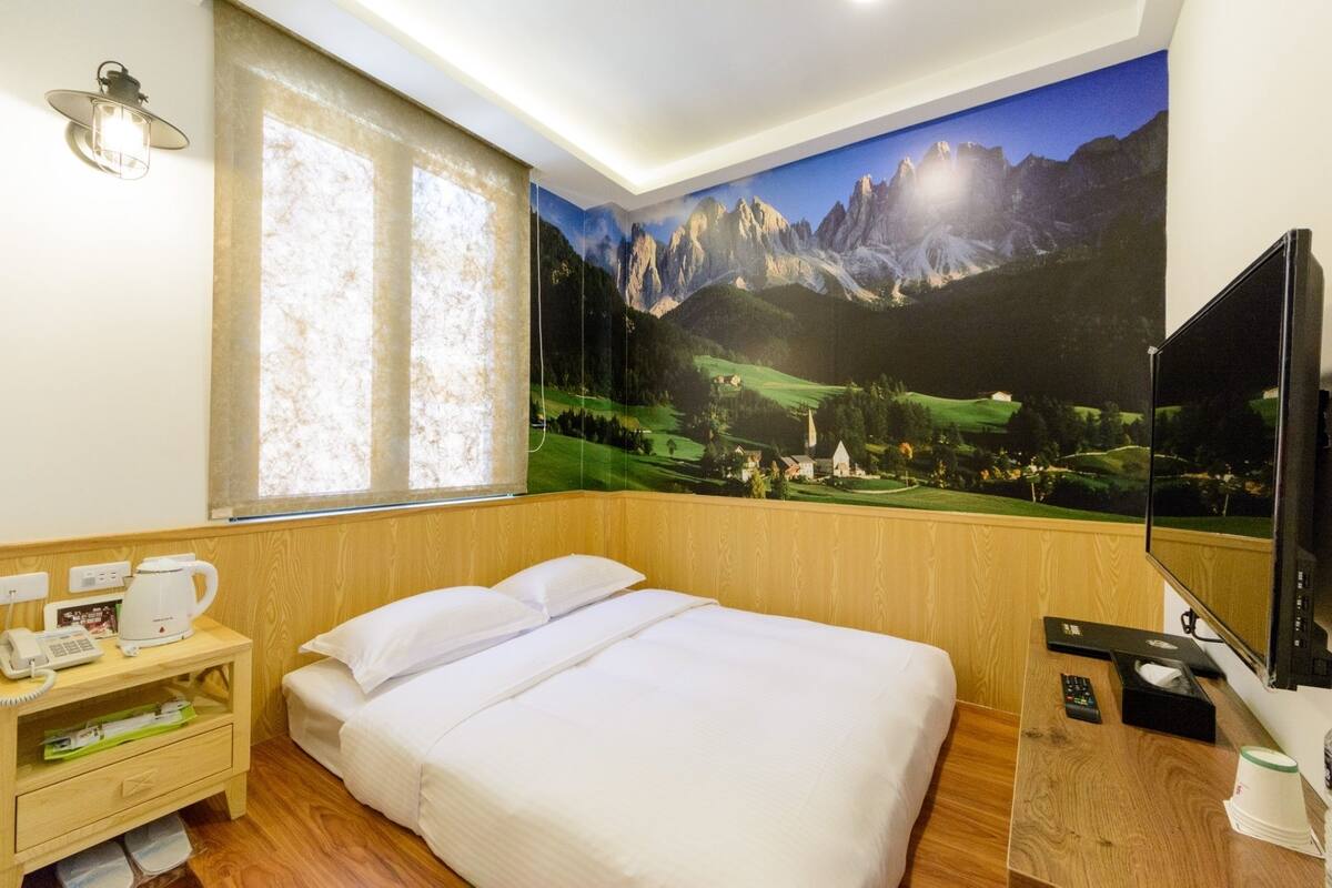 Une chambre d’hôtel avec un lit, une table de chevet sur laquelle se trouve une machine à café, une télévision et une grande fresque murale représentant un paysage de montagne.