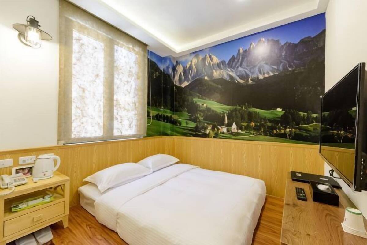 Une chambre d’hôtel comprenant un lit, une table de chevet en bois avec une machine à café, un téléviseur à écran plat et une grande fresque murale représentant un paysage de montagne.