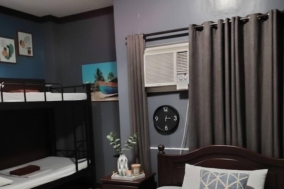 Habitación de hotel con una cama individual, una cama nido más pequeña, una mesita de noche con un reloj y una ventana con cortinas.