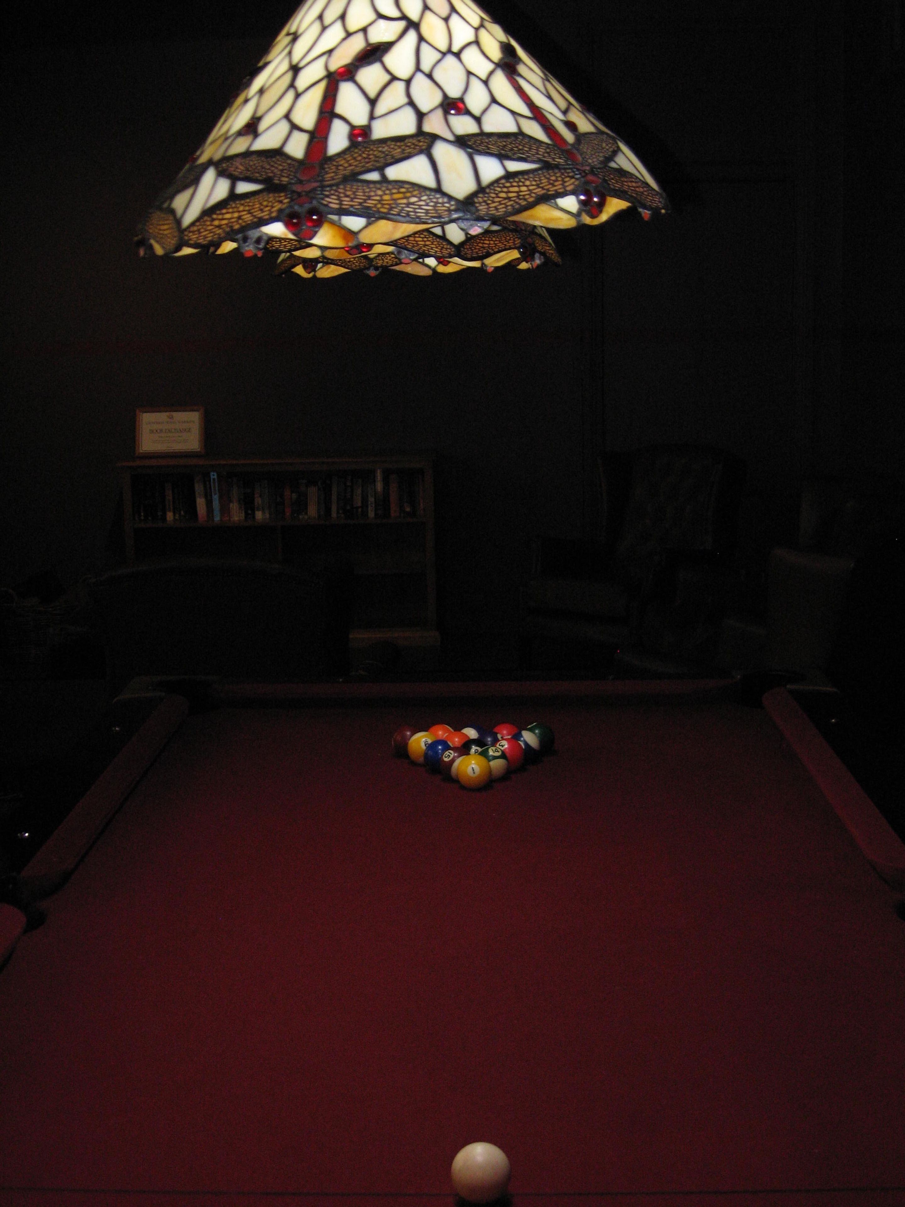 A billiards table with balls and a cue stick, a bookshelf, and a stained glass lamp.