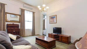 A living room with a brown sofa, a wooden coffee table, a TV on a wooden stand, a chandelier, and a large window with curtains. - Criterion Hotel Warwick (Warwick)