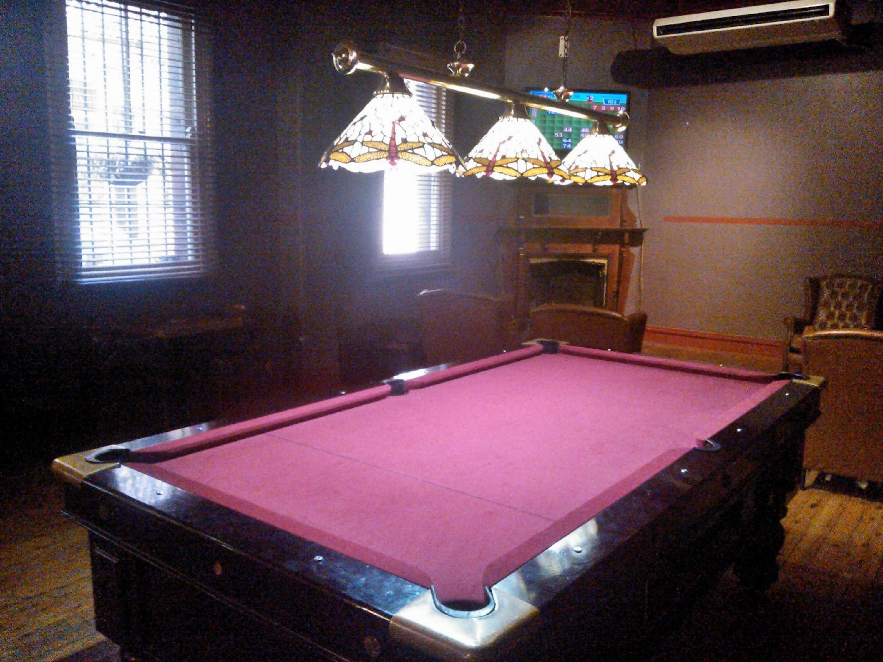 Une table de billard recouverte d’un feutre rose, un lustre orné de lampes en vitrail et une fenêtre avec des stores.