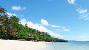 A beach with white sand, clear blue water, and lush green trees. There are thatched-roof huts along the shore. - The Stopover Hostel Mactan (Lapu-Lapu)