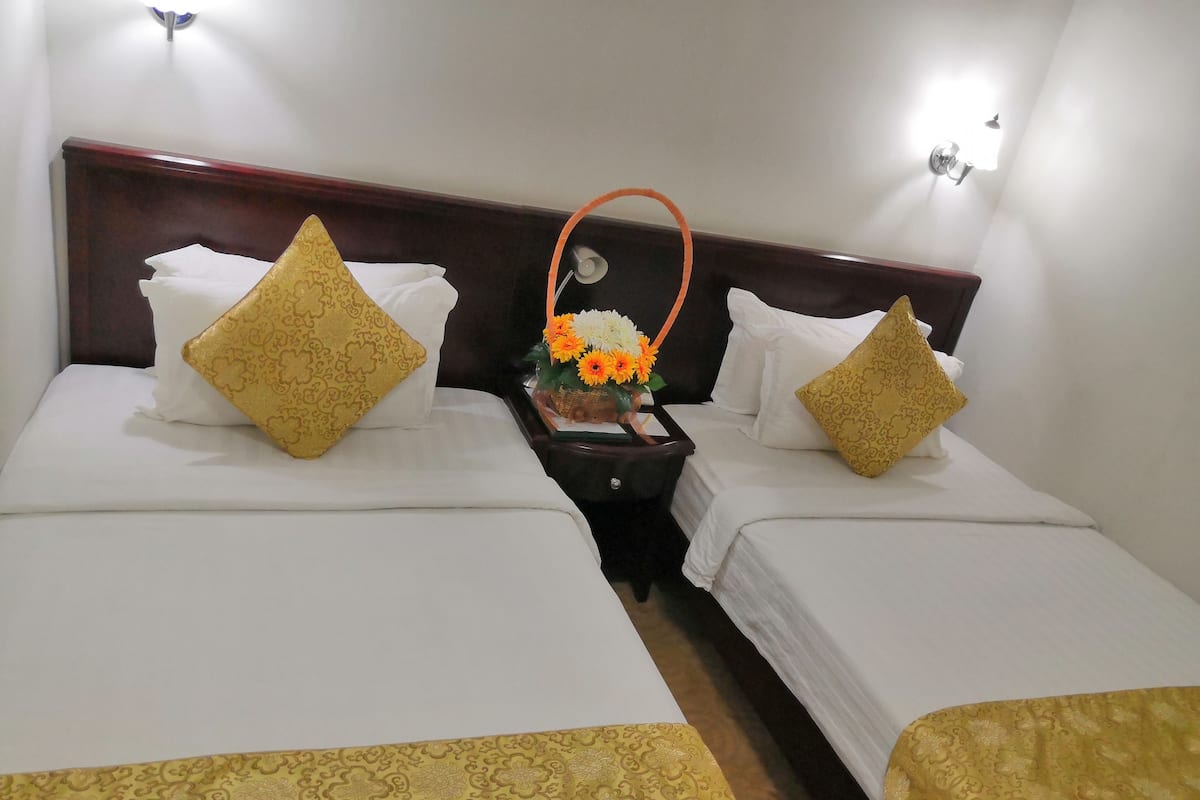 A hotel room with two beds, each with white linens and a gold-patterned bedspread. There is a nightstand between the beds with a basket of flowers and a lamp.
