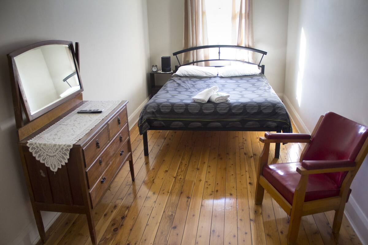 A bedroom with a wooden dresser, a red chair, a bed with a patterned bedspread, and a bedside table with a clock and lamp.
