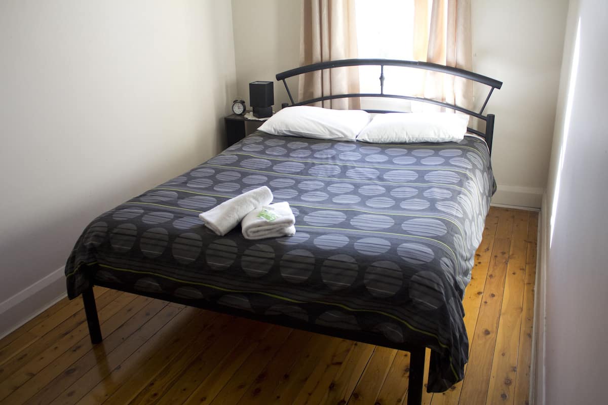 A neatly made bed with a patterned bedspread, a bedside table with a clock, and a window with curtains.