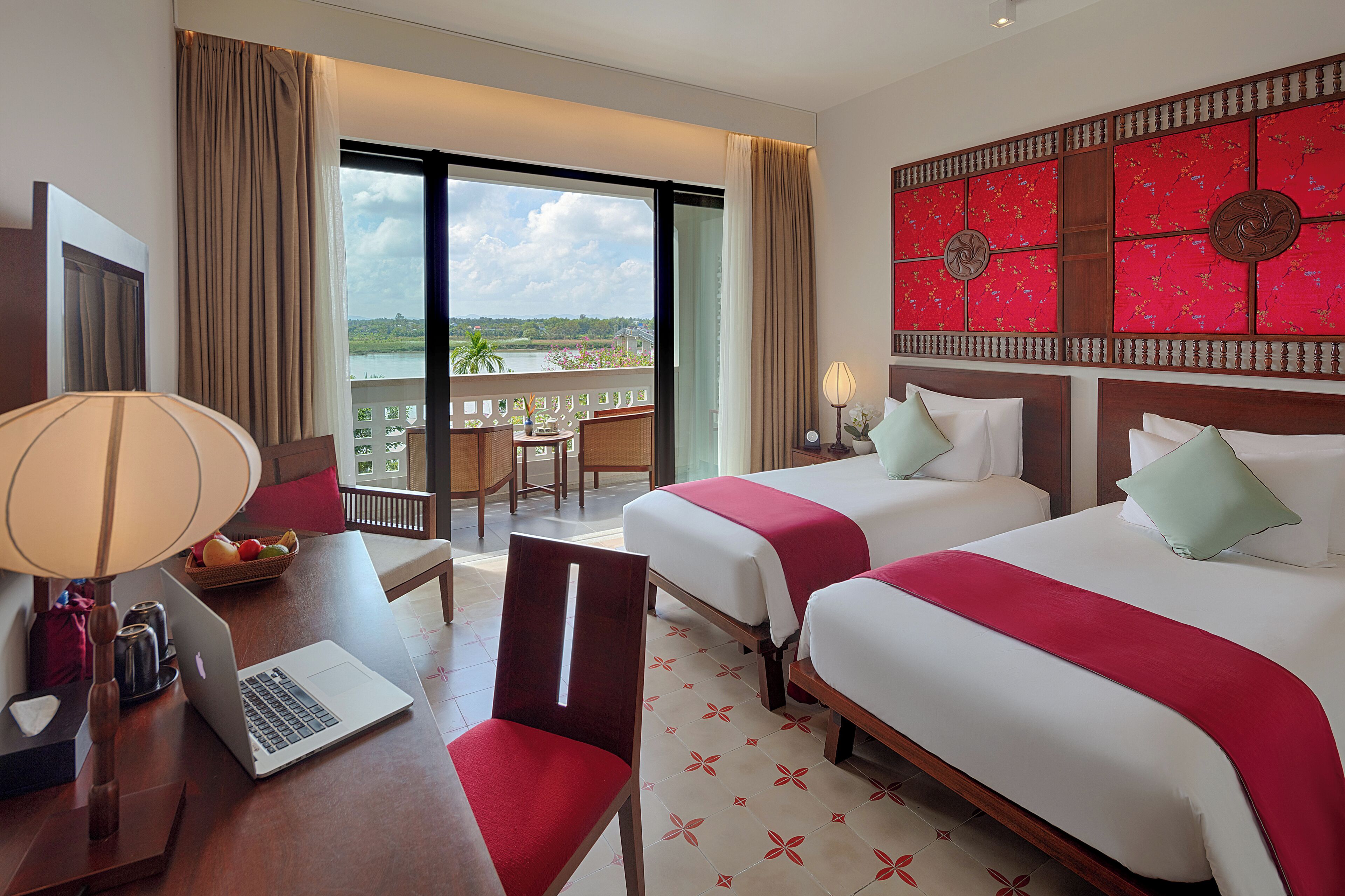 A hotel room with two beds, a desk with a laptop, a chair, a table with fruit, and a balcony with a view.