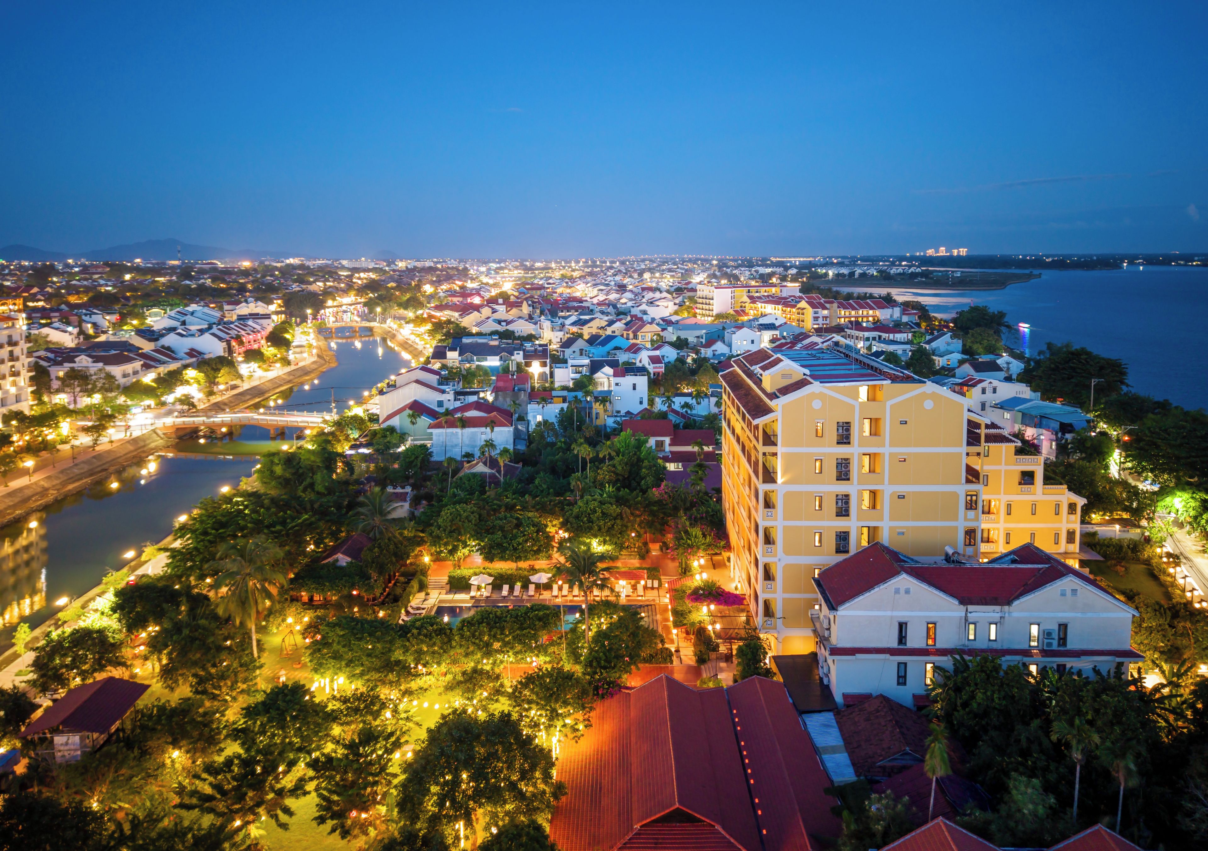A coastal cityscape at dusk with illuminated buildings, a river, and a large hotel with multiple floors and balconies.