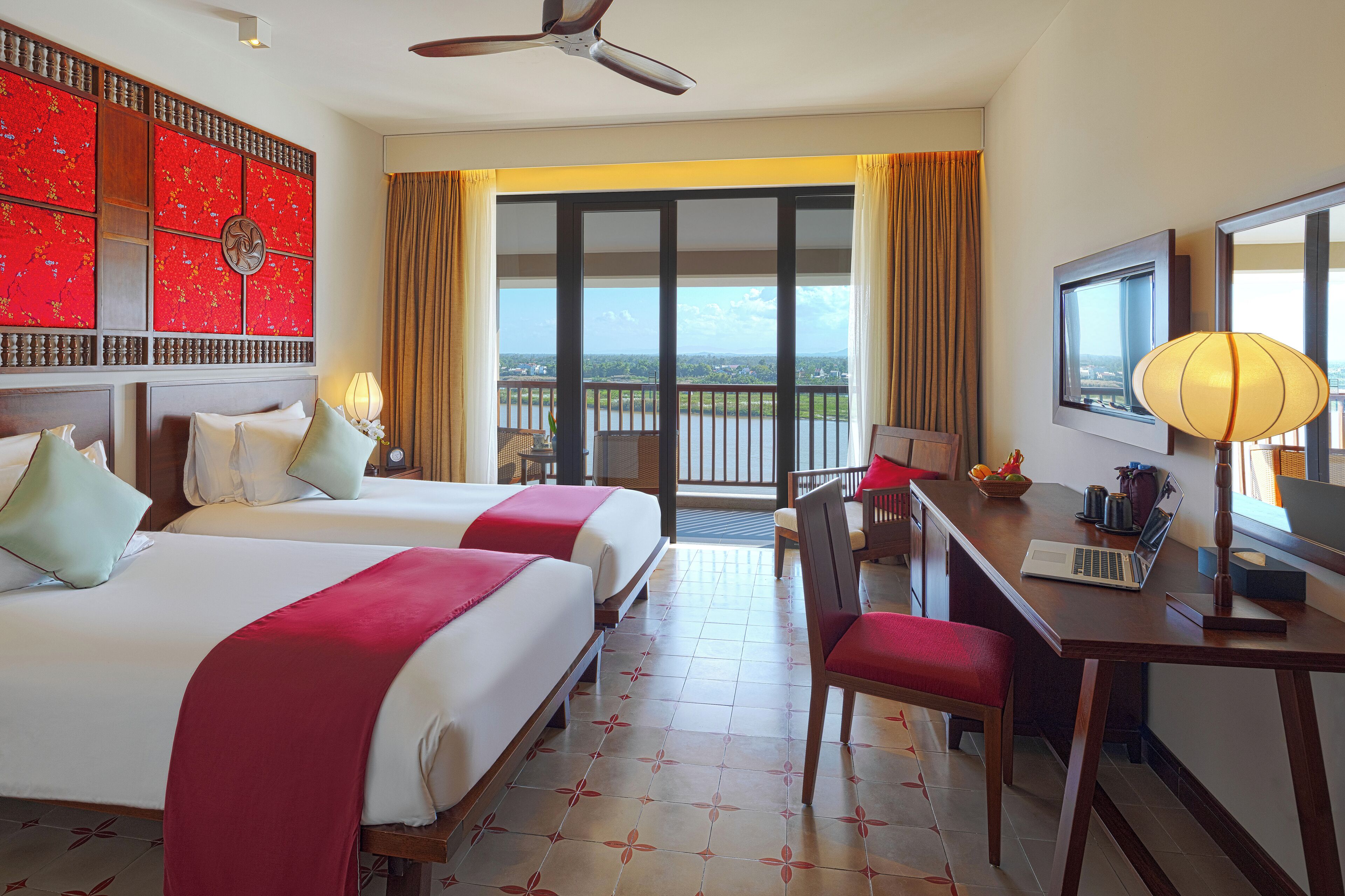 A hotel room with two beds, a desk with a laptop, a television, and a balcony with a view.