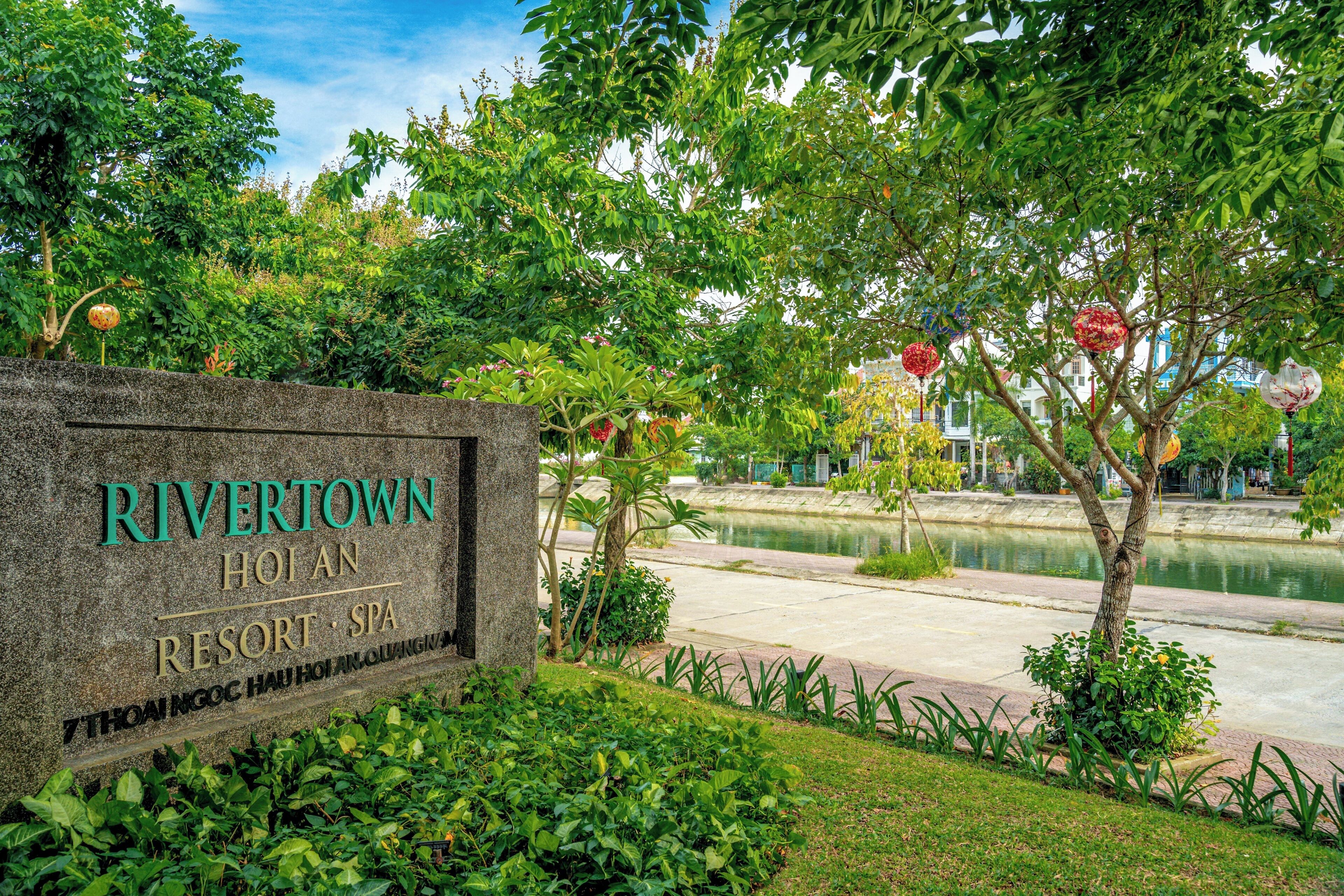 Un letrero de piedra con el texto 'RIVERTOWN HOTEL RESORT SPA', rodeado de exuberante follaje y una masa de agua.