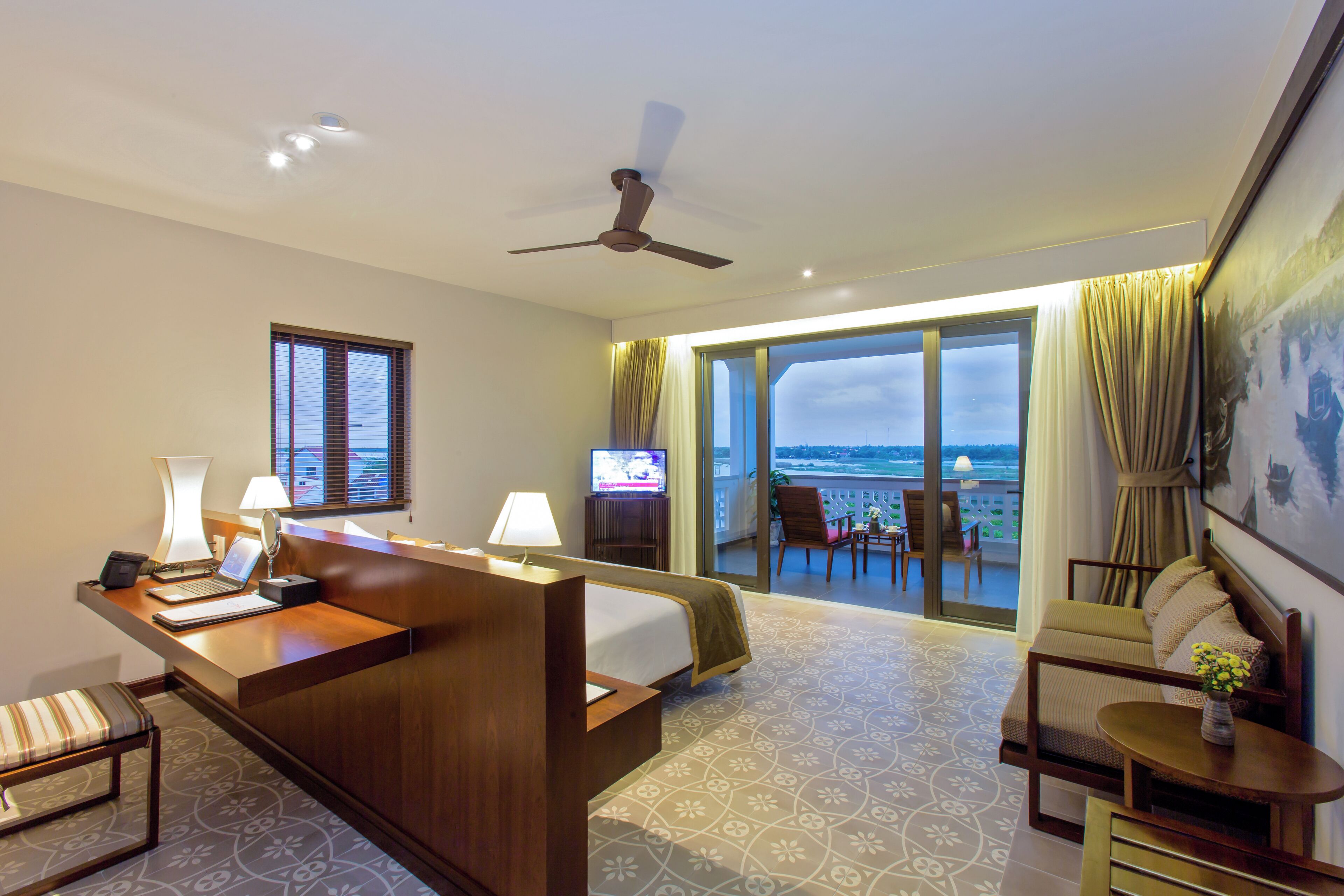 A hotel room with a bed, desk, and seating area, a television, and a balcony with a view.