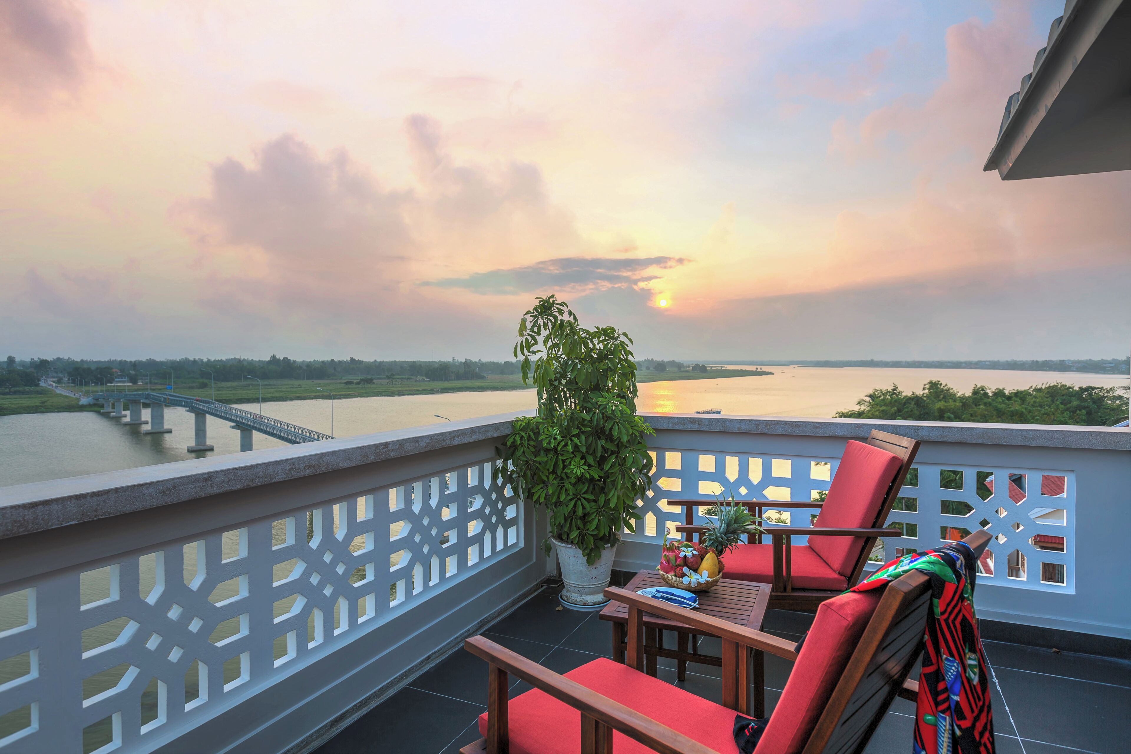 A balcony with a view of a river, a bridge, and a sunset. There are red chairs and a table with a plant and fruit basket.