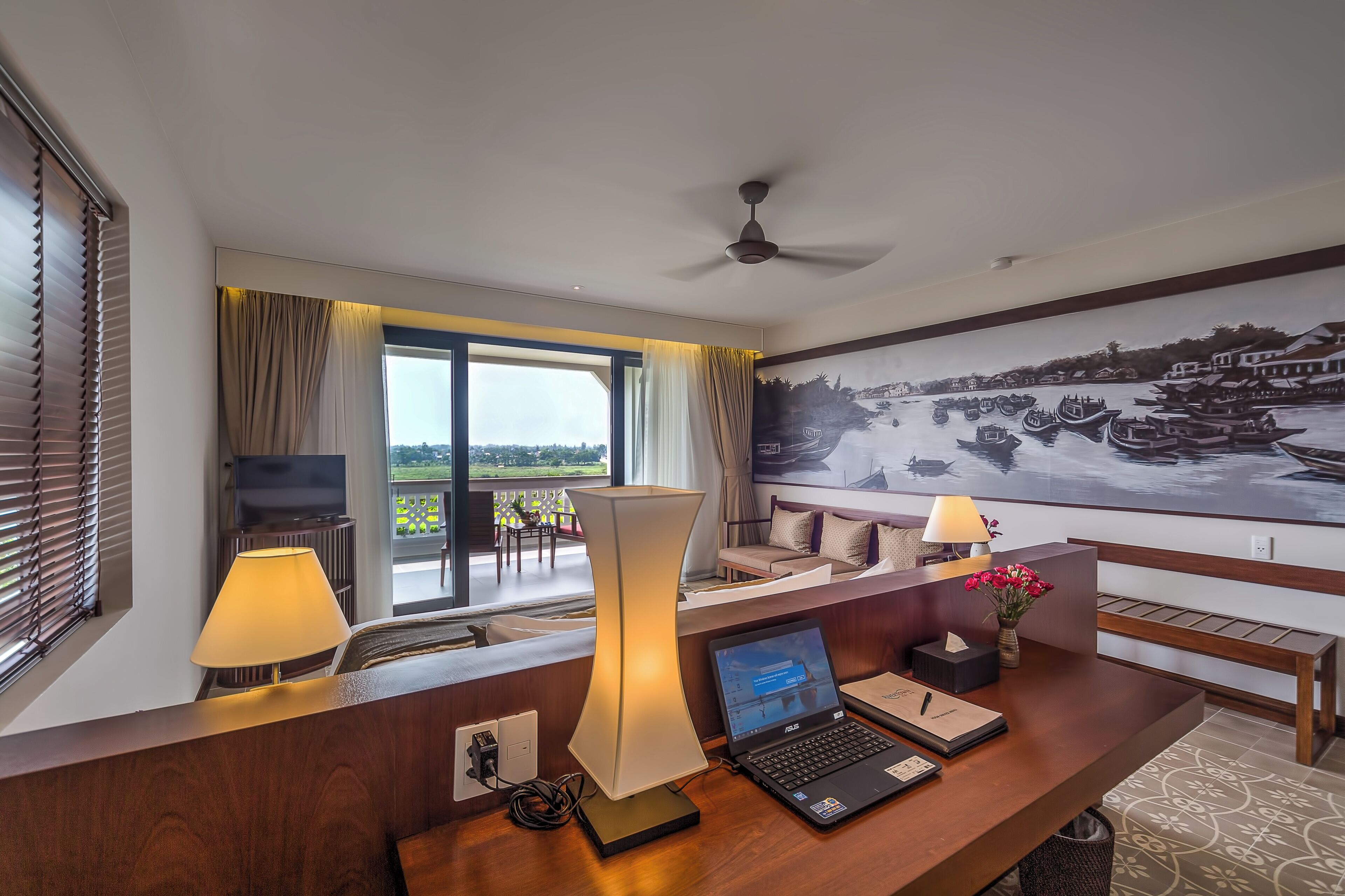 Una camera d'albergo con scrivania, laptop, lampada, televisore e una grande finestra con tende.