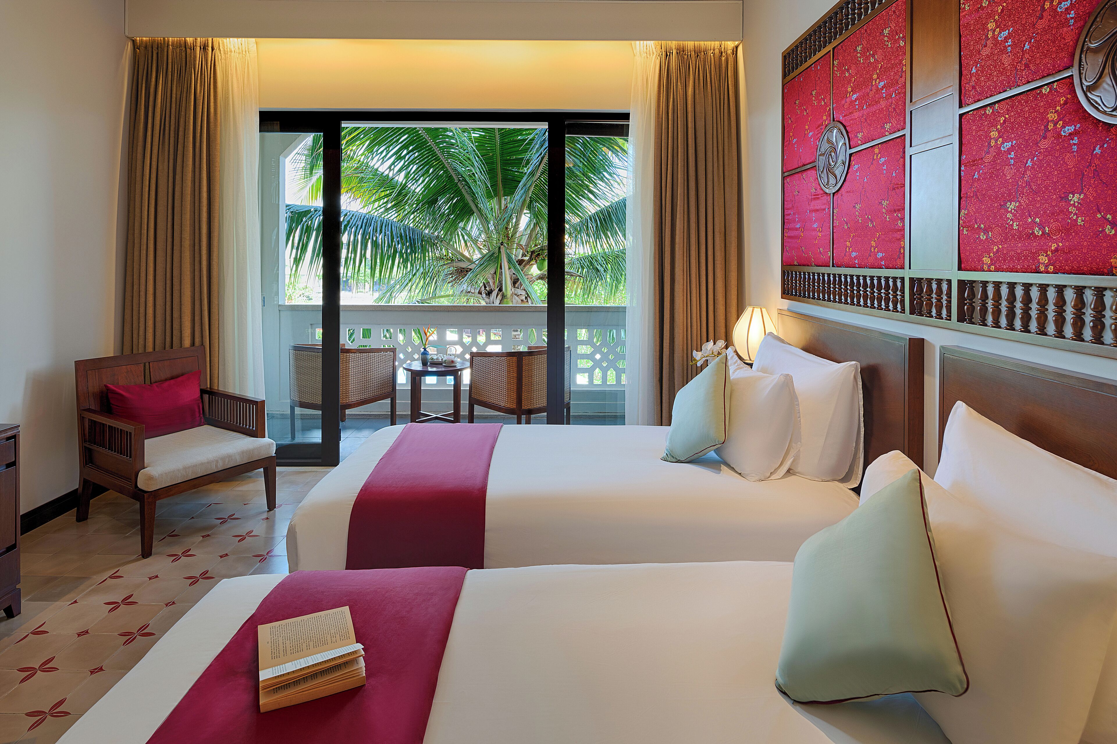 A hotel room with two beds, a sitting area with a chair and table, and a view of palm trees through large windows.