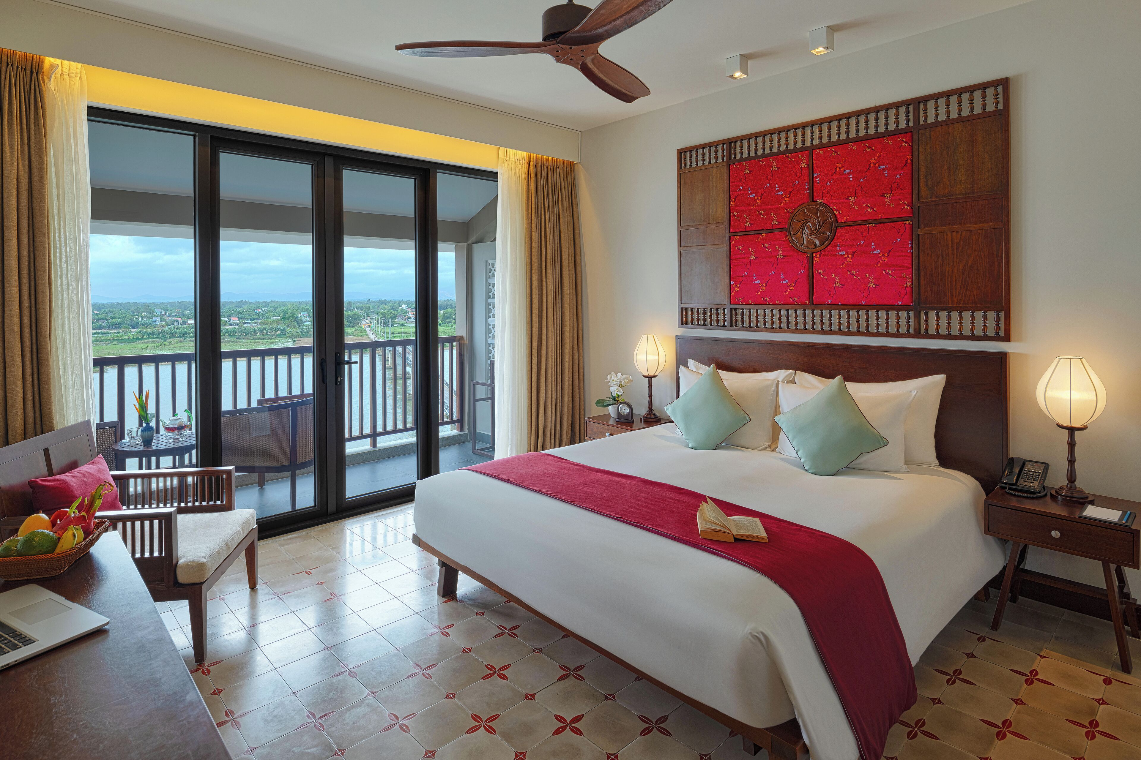 A hotel room with a bed, a desk with a laptop, a chair, a balcony with a view, and a ceiling fan.