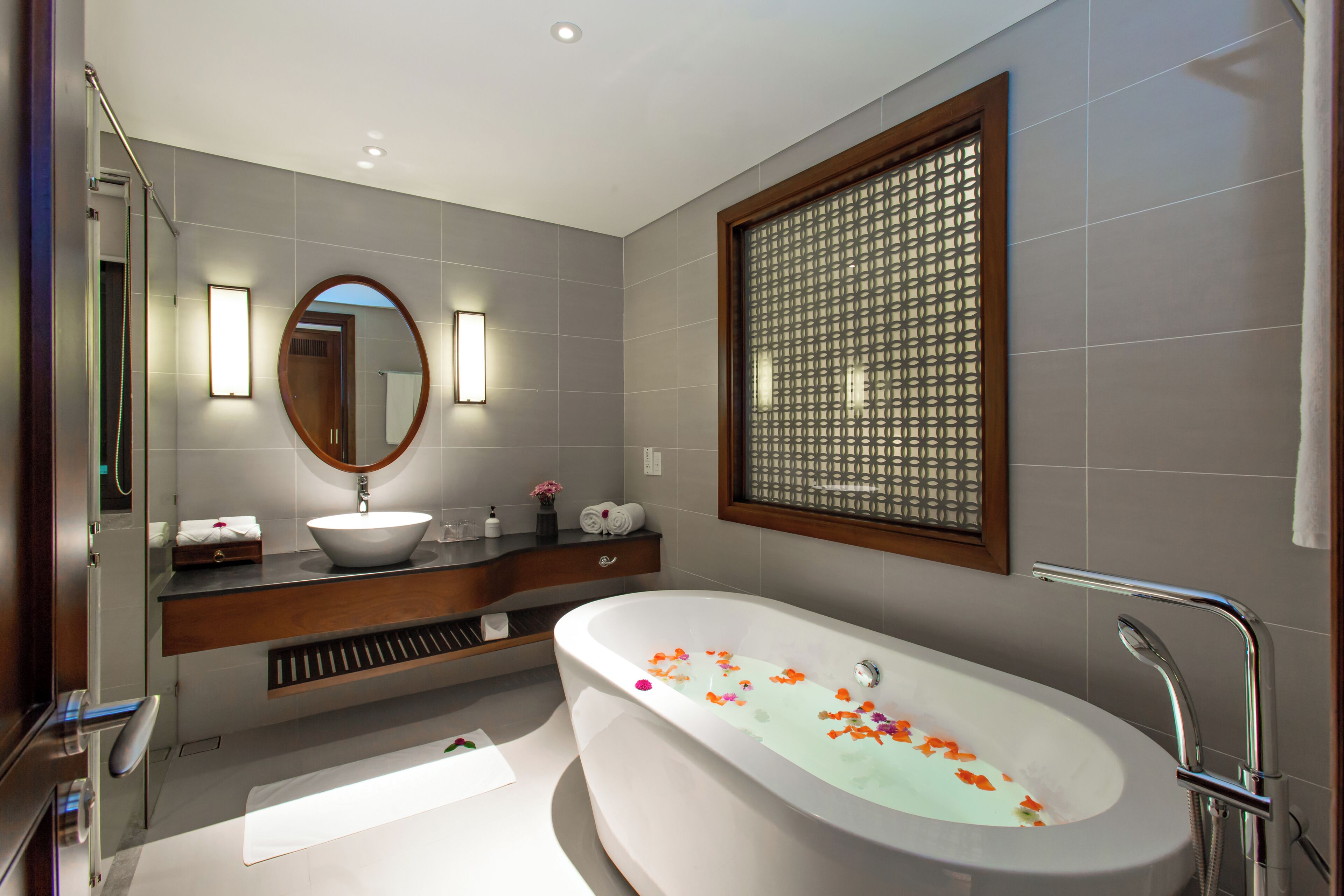 A modern bathroom with a freestanding bathtub, a large mirror, and a window with a decorative screen.