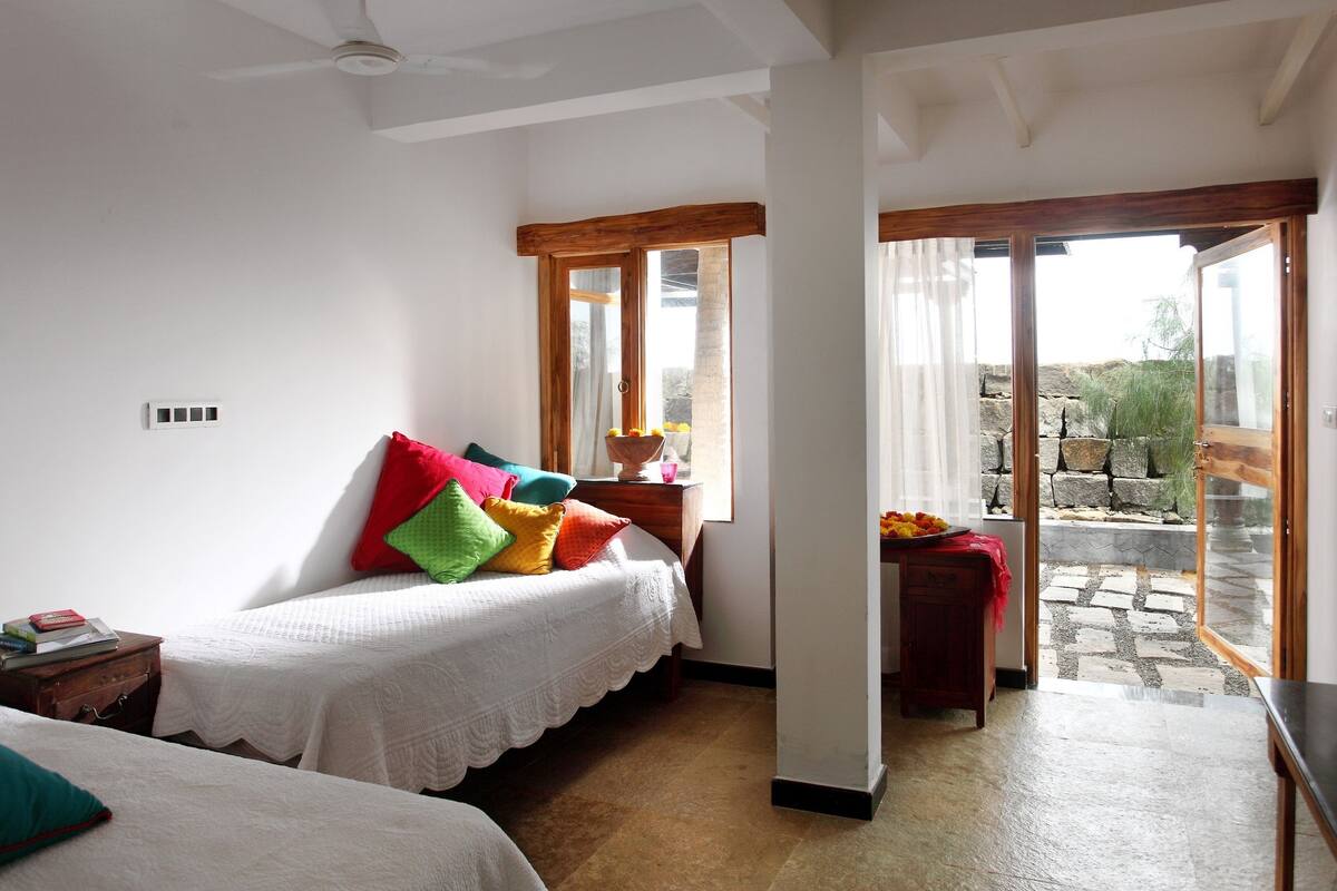 A bedroom with a bed, a nightstand, a window with a view of a garden, and a door leading outside.