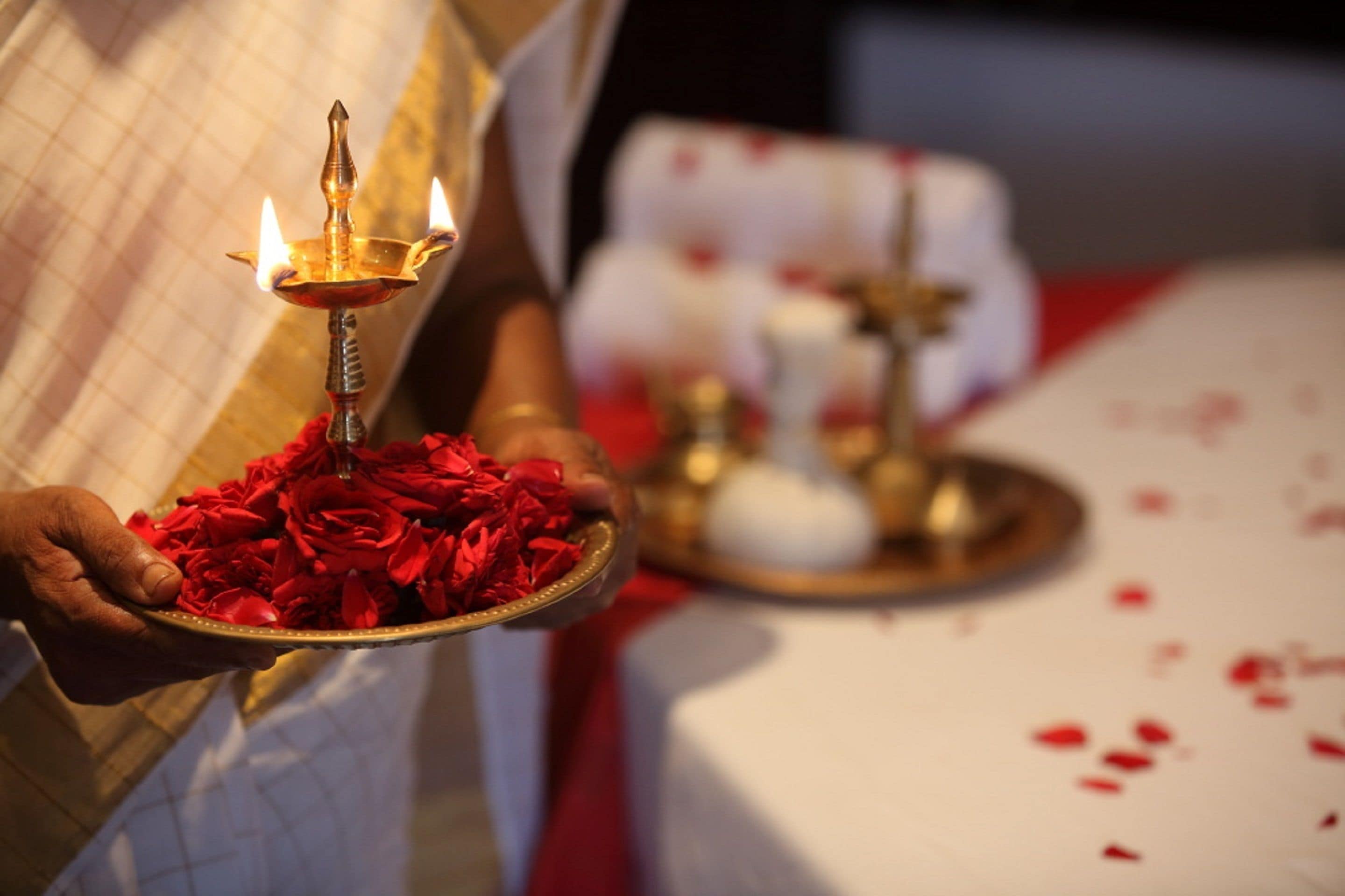A person holding a tray with a lit oil lamp and red flowers.