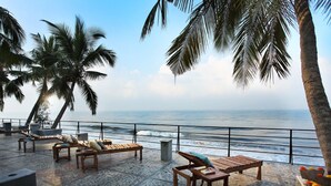 A beachside lounge with wooden furniture and palm trees, overlooking the ocean. - 36 Palms Boutique Retreat (Kochi)
