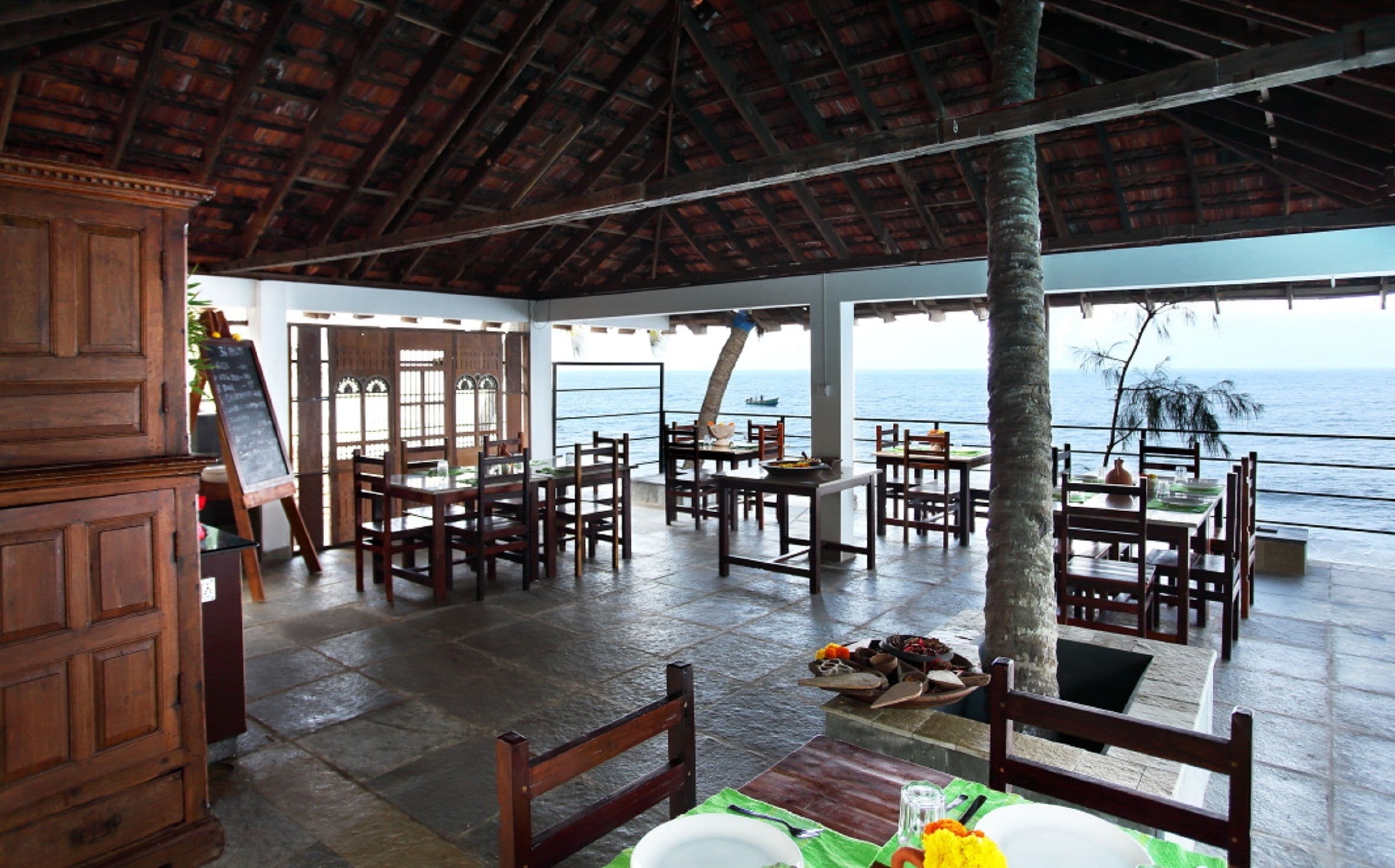 An open-air dining area with wooden tables and chairs, a view of the ocean, and a thatched roof.