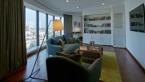 Una sala de estar moderna con sofá, mesa de centro y estantería, ventanas amplias con cortinas y vista a la ciudad.