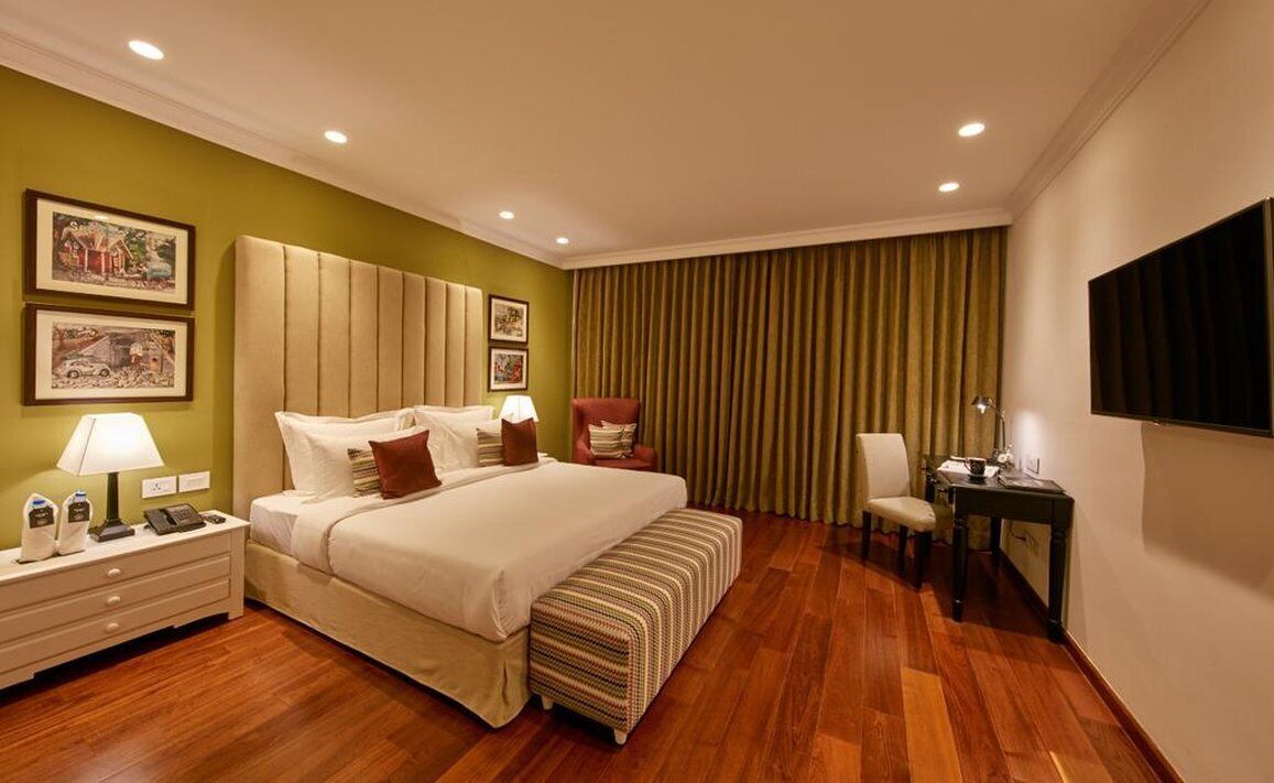 Un cuarto de hotel con una cama grande, una mesita de noche con lámpara, una silla, un escritorio con silla, y un televisor montado en la pared.