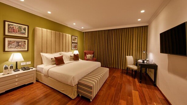 A hotel room with a large bed, a bedside table with a lamp, a chair, a desk with a chair, and a TV mounted on the wall. - The Waverly Hotel & Residences (Bengaluru)