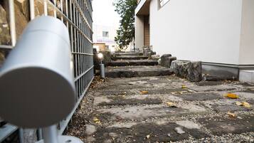 Un étroit sentier de pierres menant à l’entrée d’un bâtiment, avec une clôture en métal et une caméra de surveillance sur le côté gauche.
