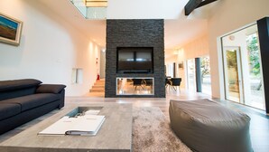 A modern living room with a stone fireplace, a large flat-screen TV, a dark sofa, and a large window offering a view of the outdoors. - La Cachette (Hakodate)