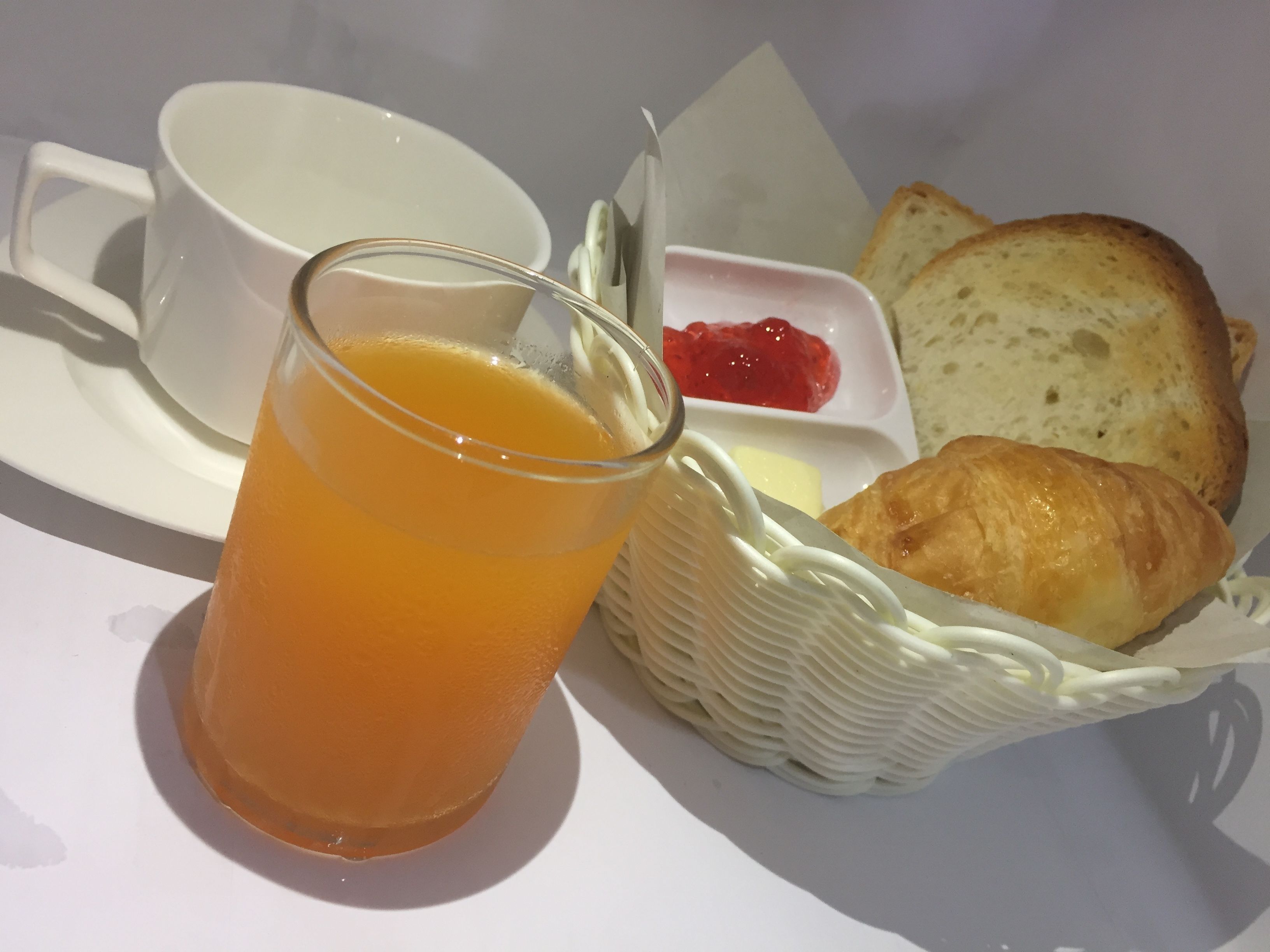 Una escena de desayuno con un vaso de jugo de naranja, una taza, una cesta con pan y un croissant, y un recipiente pequeño de mermelada.