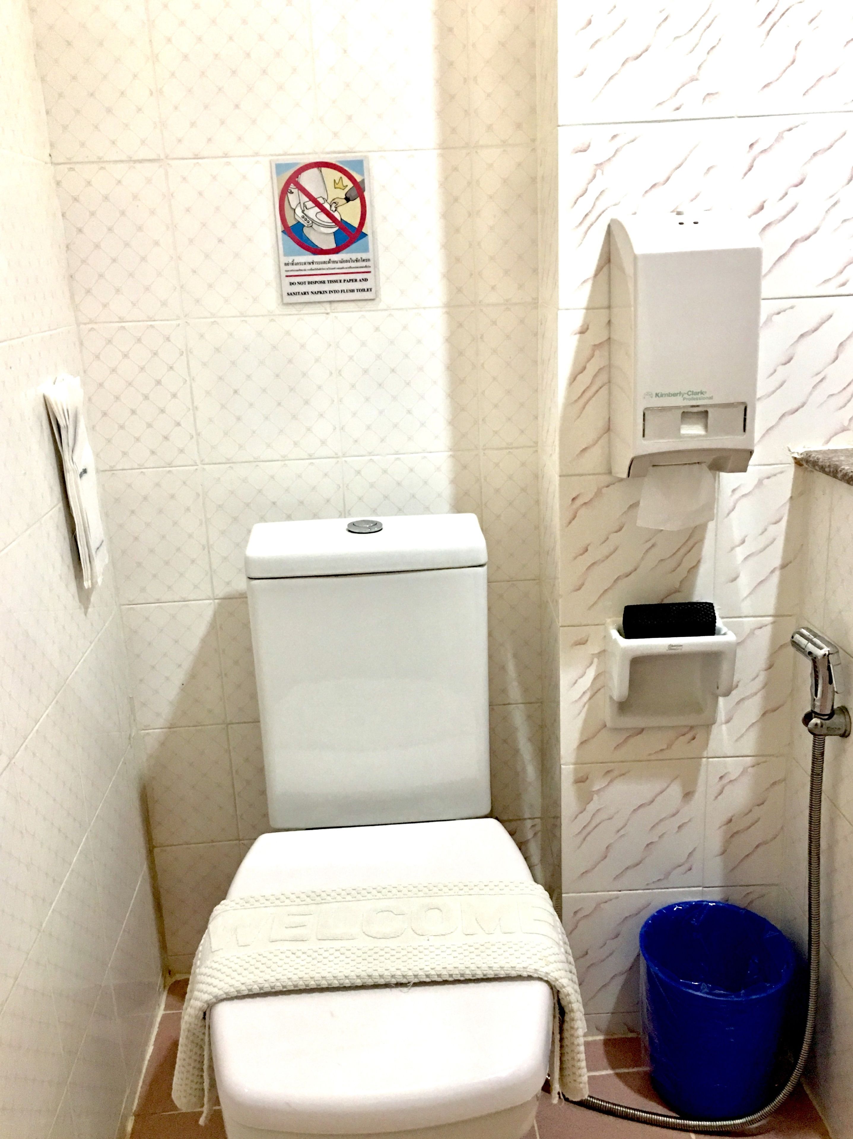 A small, compact bathroom with a toilet, a hand dryer, a paper towel dispenser, and a blue trash bin.