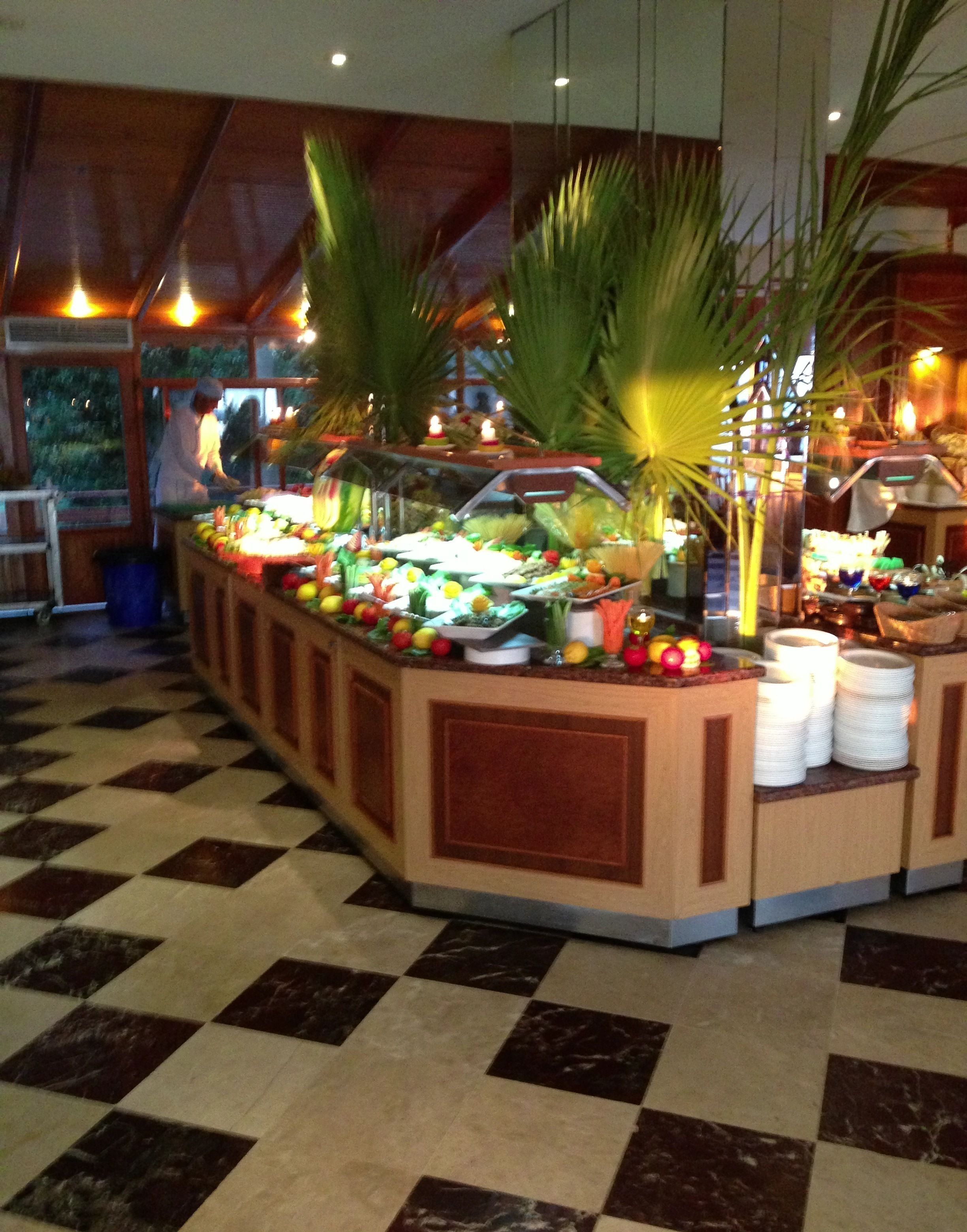 Un espace buffet doté d’un sol à carreaux, d’un comptoir central où sont présentés différents plats, et agrémenté de plantes tropicales.