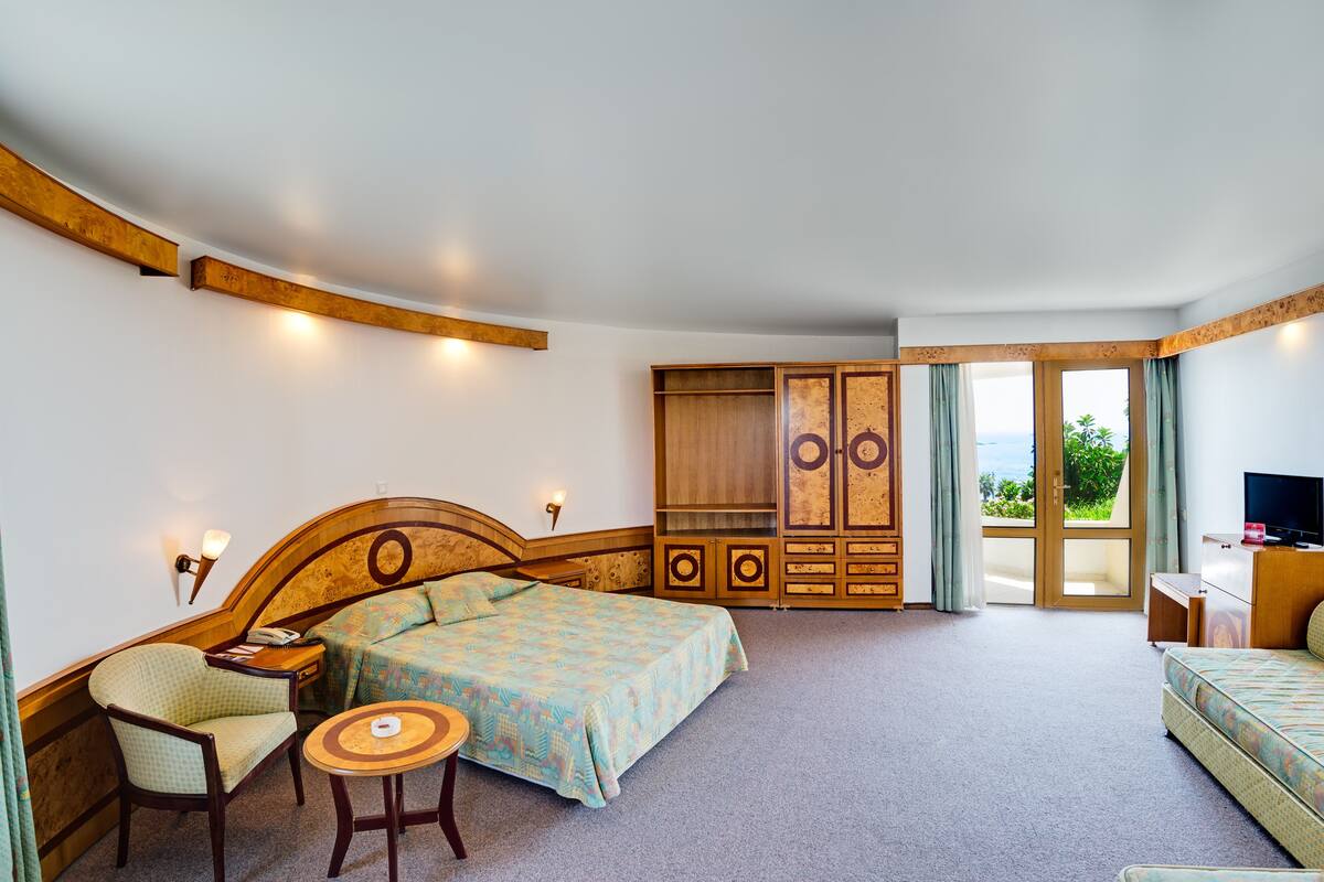 A bedroom with a bed, a chair, a small table, a wardrobe, and a TV.