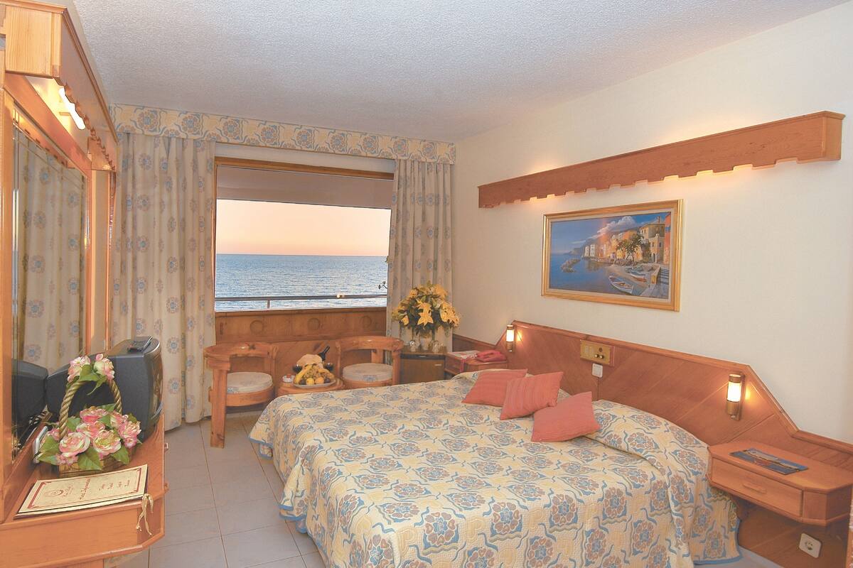A bedroom with a bed, a desk with a computer, a chair, a window with a view of the sea, and a painting on the wall.