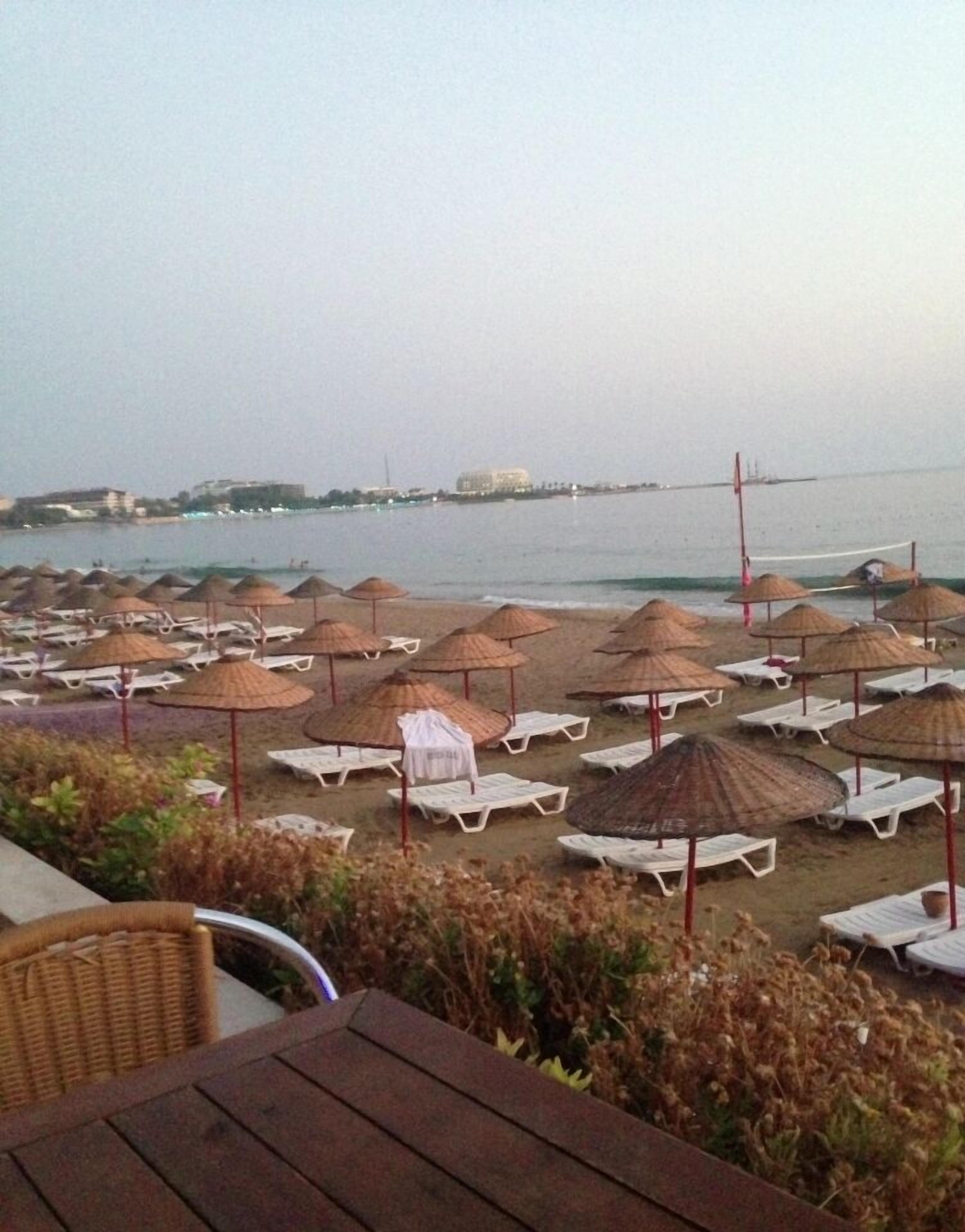 A beach with rows of sun loungers and thatched umbrellas, a view of the sea, and buildings in the distance.