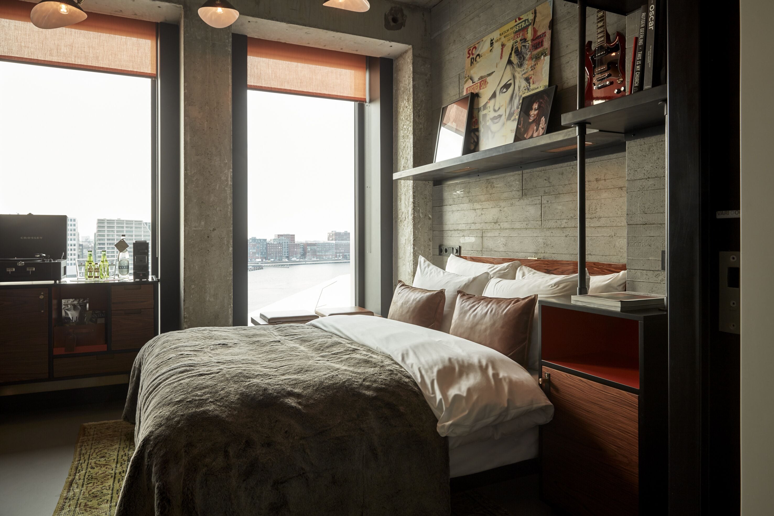 A modern bedroom with a large bed, a wooden dresser, a bookshelf, and a city view through large windows.