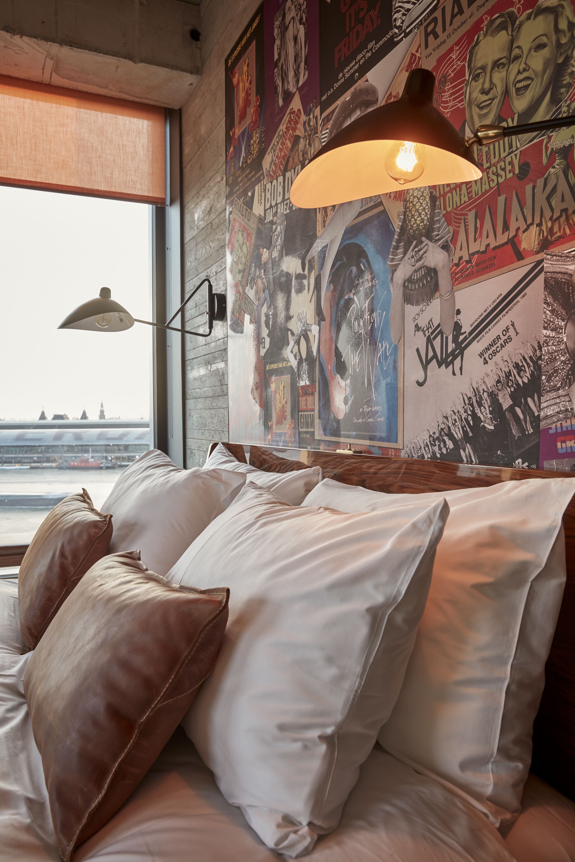A bed with white and brown pillows, a hanging lamp, and a poster-covered wall.