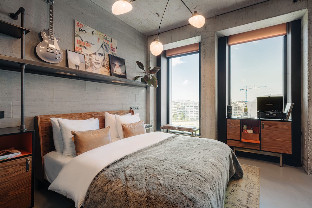 A modern hotel room with a large bed, a wooden headboard, a desk with a computer, and a large window with a city view.
