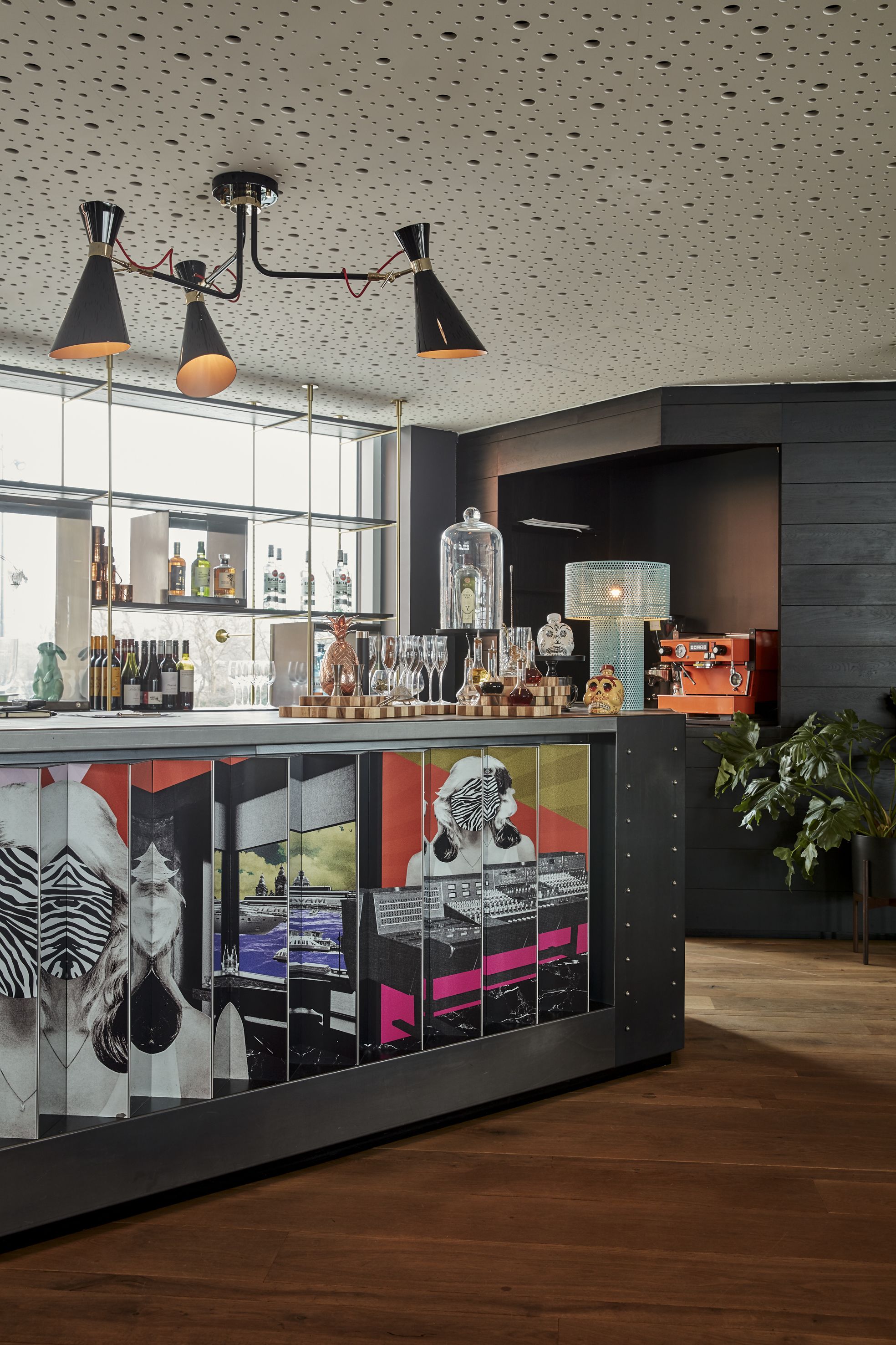 Un espace bar moderne doté d’un plafond perforé, d’un lustre central et d’un comptoir où sont exposées diverses bouteilles et différents types de verres.