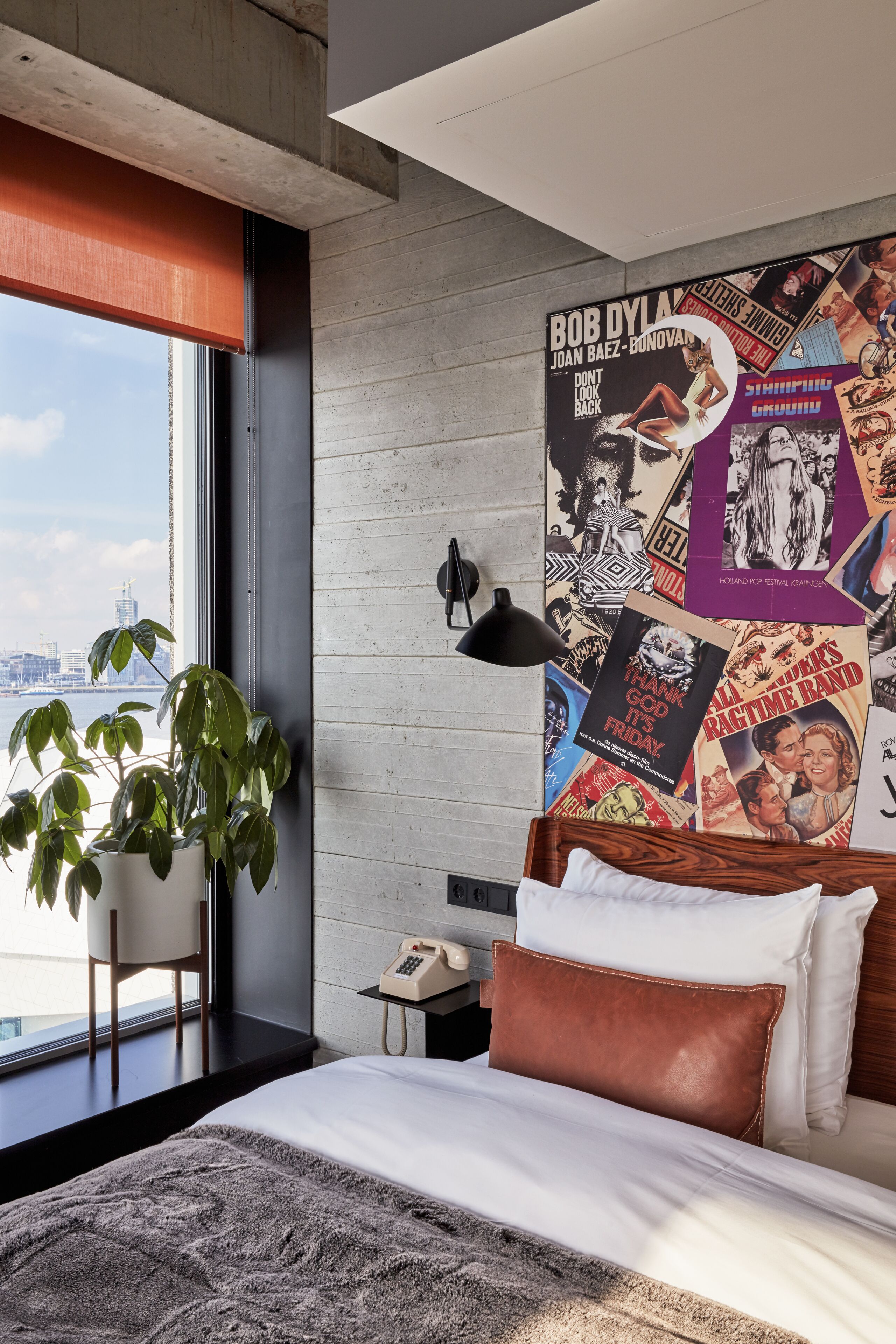 A modern hotel room with a large bed, a city view, a poster on the wall, and a plant by the window.