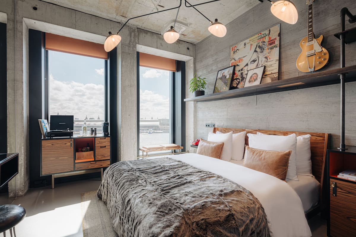 A modern hotel room with a large bed, a desk with a computer, and a view of the city through large windows.